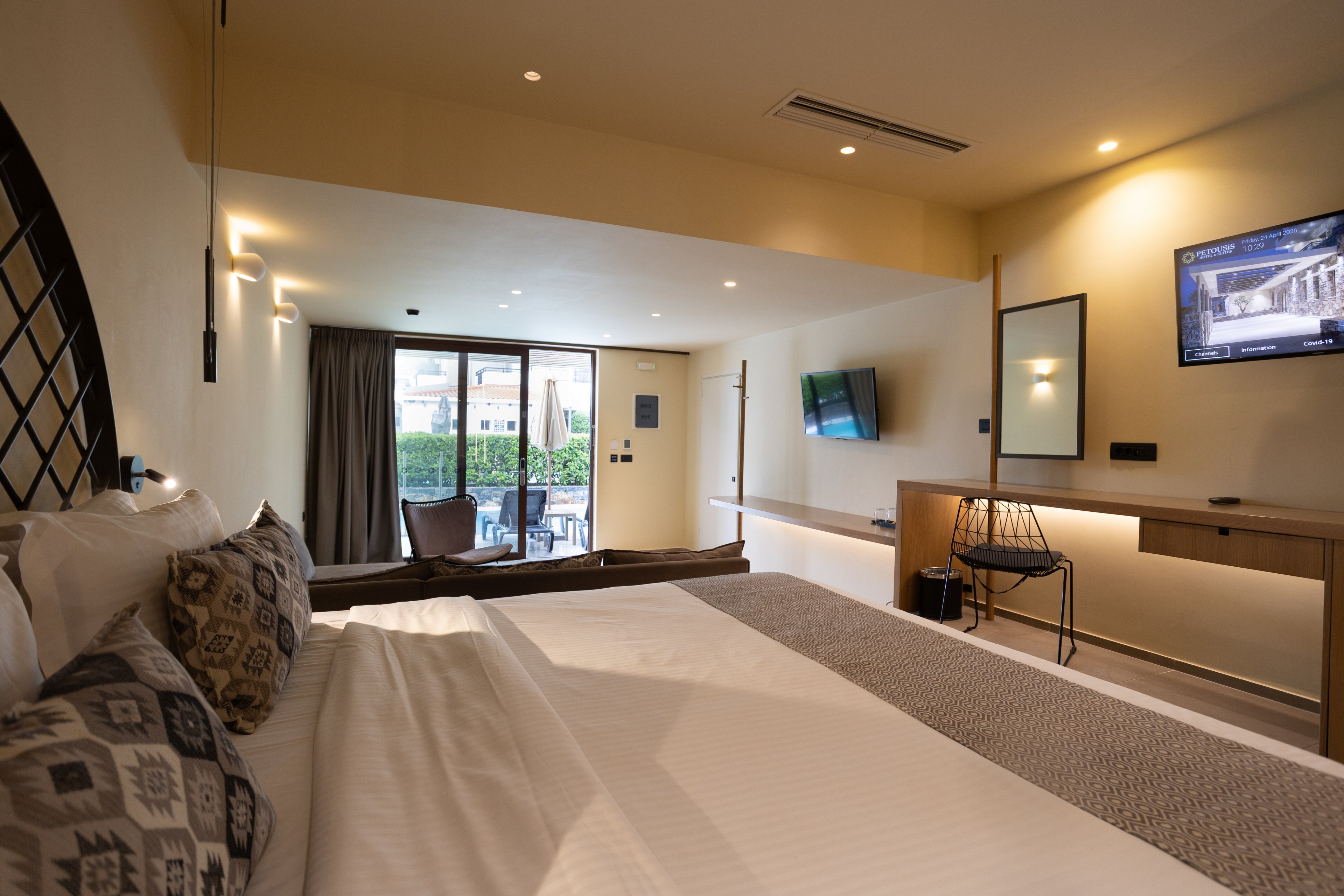 A spacious modern hotel room features a large neatly made bed in the foreground with patterned pillows. The room includes a wall-mounted television, a long wooden desk with a chair, and warm recessed lighting. Sliding glass doors open to an outdoor terrace area with greenery and additional seating. Neutral tones and soft lighting create a calm, inviting atmosphere suitable for relaxation or business travel.