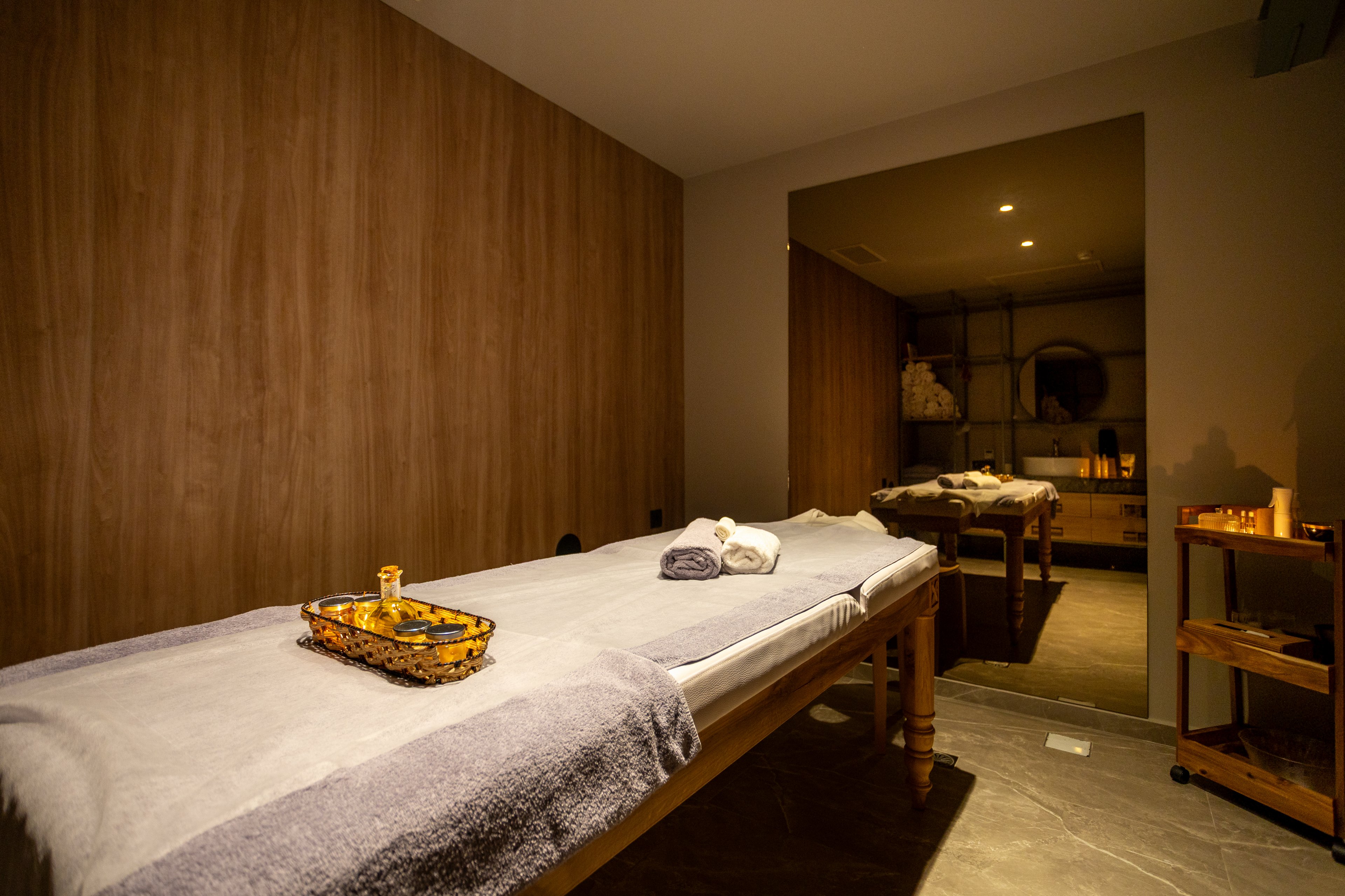 A neatly prepared massage table is set in a cozy spa treatment room. Soft towels and a small tray of spa accessories rest on the bed, ready for a client. Warm ambient lighting and wooden wall panels create a relaxing atmosphere. In the background, shelves with folded linens and decor enhance the serene, wellness-focused setting.