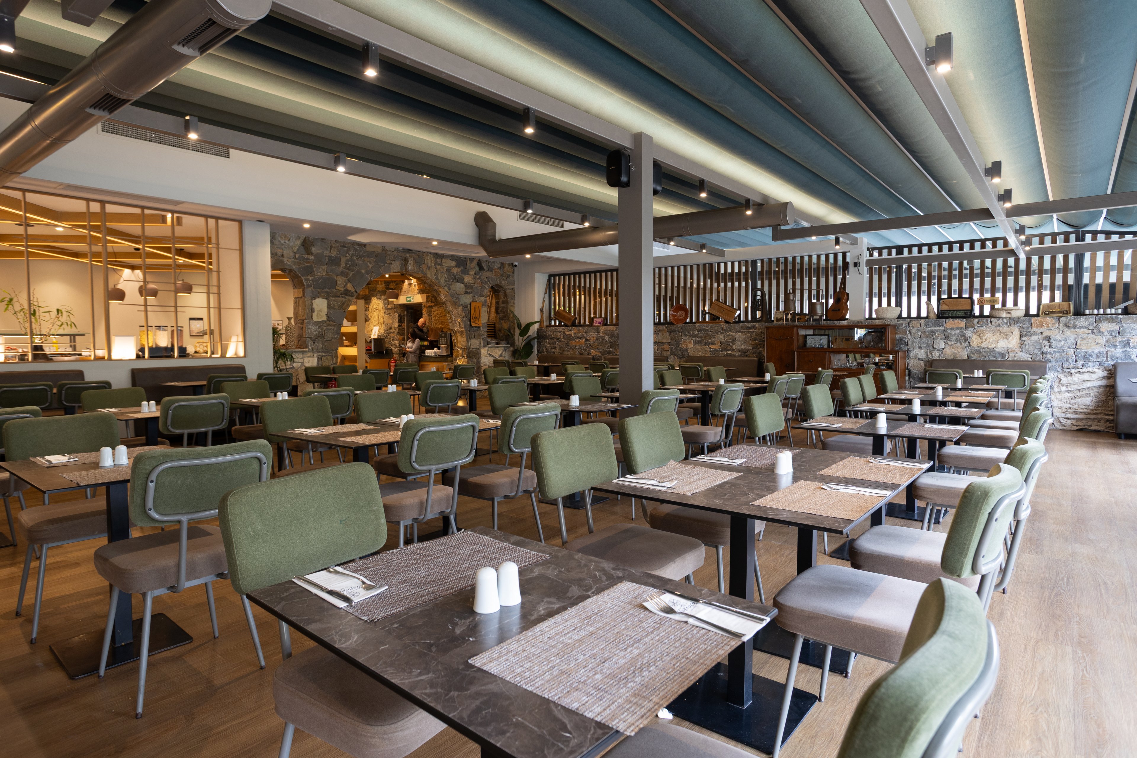 A contemporary restaurant interior features rows of neatly arranged tables and green upholstered chairs. The space is empty of guests, highlighting the clean table settings with placemats, cutlery, and salt and pepper shakers. Overhead, layered ceiling panels and track lighting create a modern atmosphere, while wooden accents and stone walls add warmth. Large windows and open partitions contribute to an airy, inviting dining environment.