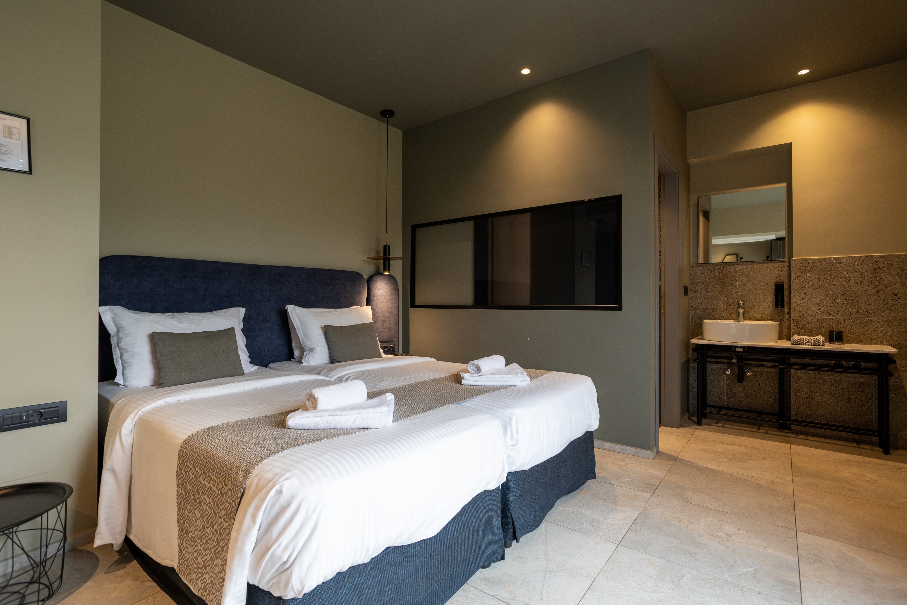 A contemporary hotel room features two neatly made twin beds with white linens and beige throws. Rolled towels are placed on each bed, adding a welcoming touch. The room has minimalist decor with neutral walls, soft lighting, and a large dark headboard. In the background, an open bathroom area with a sink and mirror is visible, enhancing the modern, open-plan design.