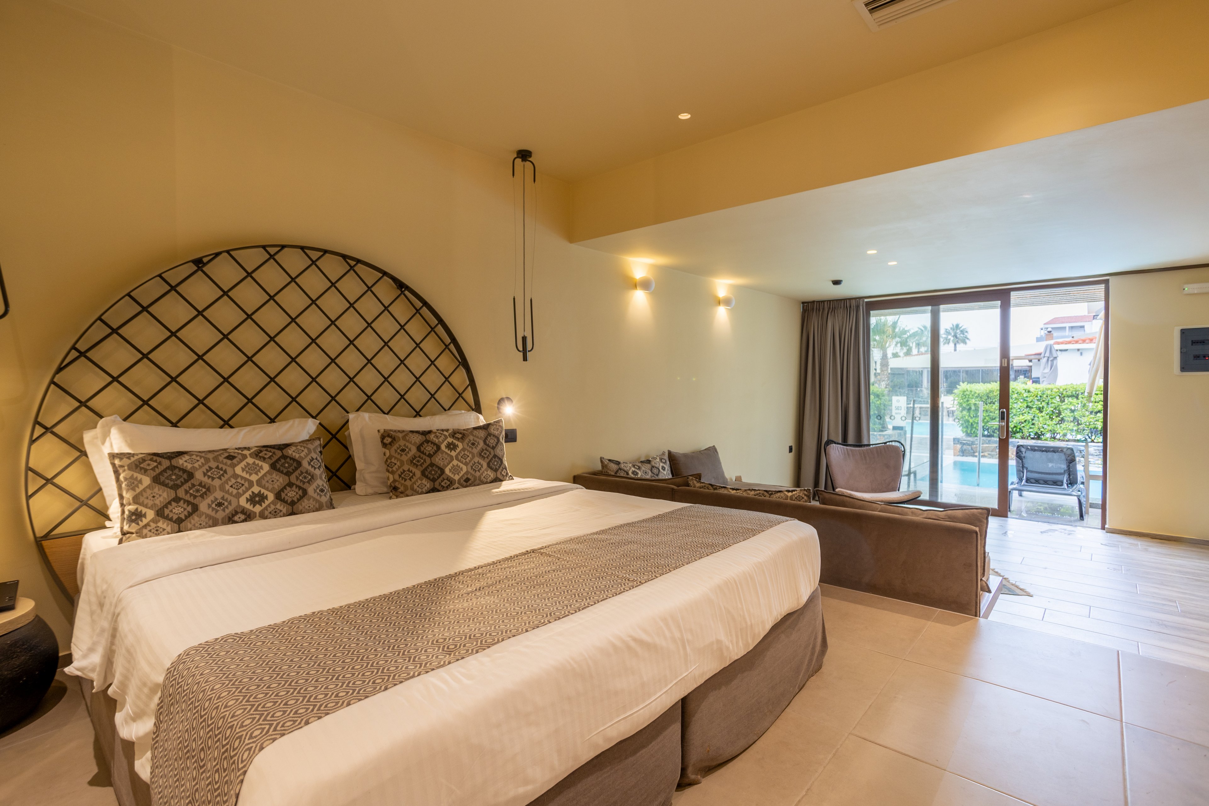 A spacious hotel room features a large bed with patterned pillows and a decorative headboard. Warm ambient lighting and neutral beige tones create a cozy, modern atmosphere. A sitting area with chairs and a table is positioned near large sliding glass doors. The doors open to a terrace with views of a pool and palm trees outside.