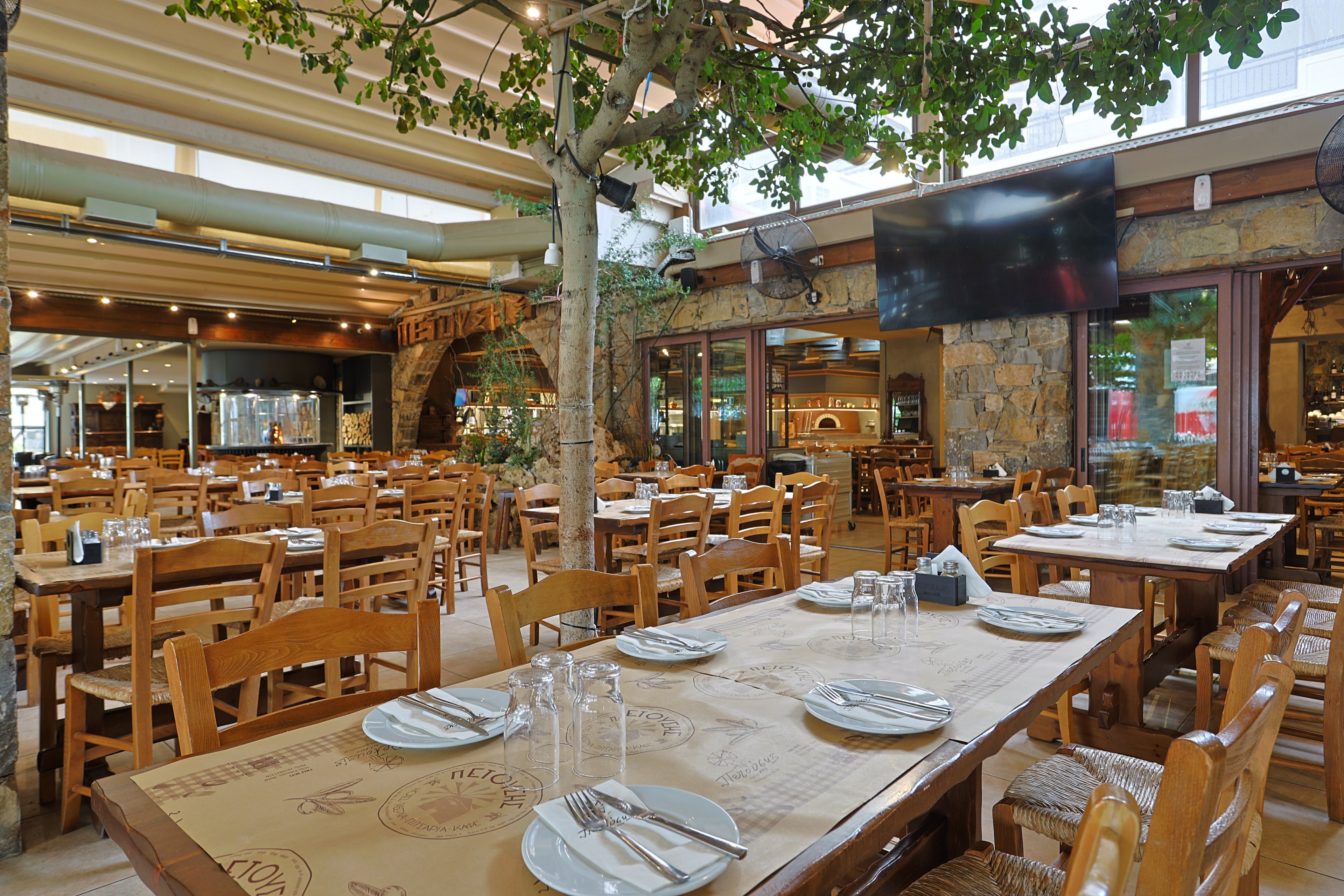 A spacious restaurant interior features neatly arranged wooden tables and chairs set with plates, glasses, and cutlery. Natural light streams in through large windows and a partially open ceiling, creating an airy atmosphere. A tree grows through the floor near the center, adding a touch of greenery to the rustic stone and wood decor. In the background, a large flat-screen television and open kitchen area are visible, suggesting a casual yet welcoming dining experience.