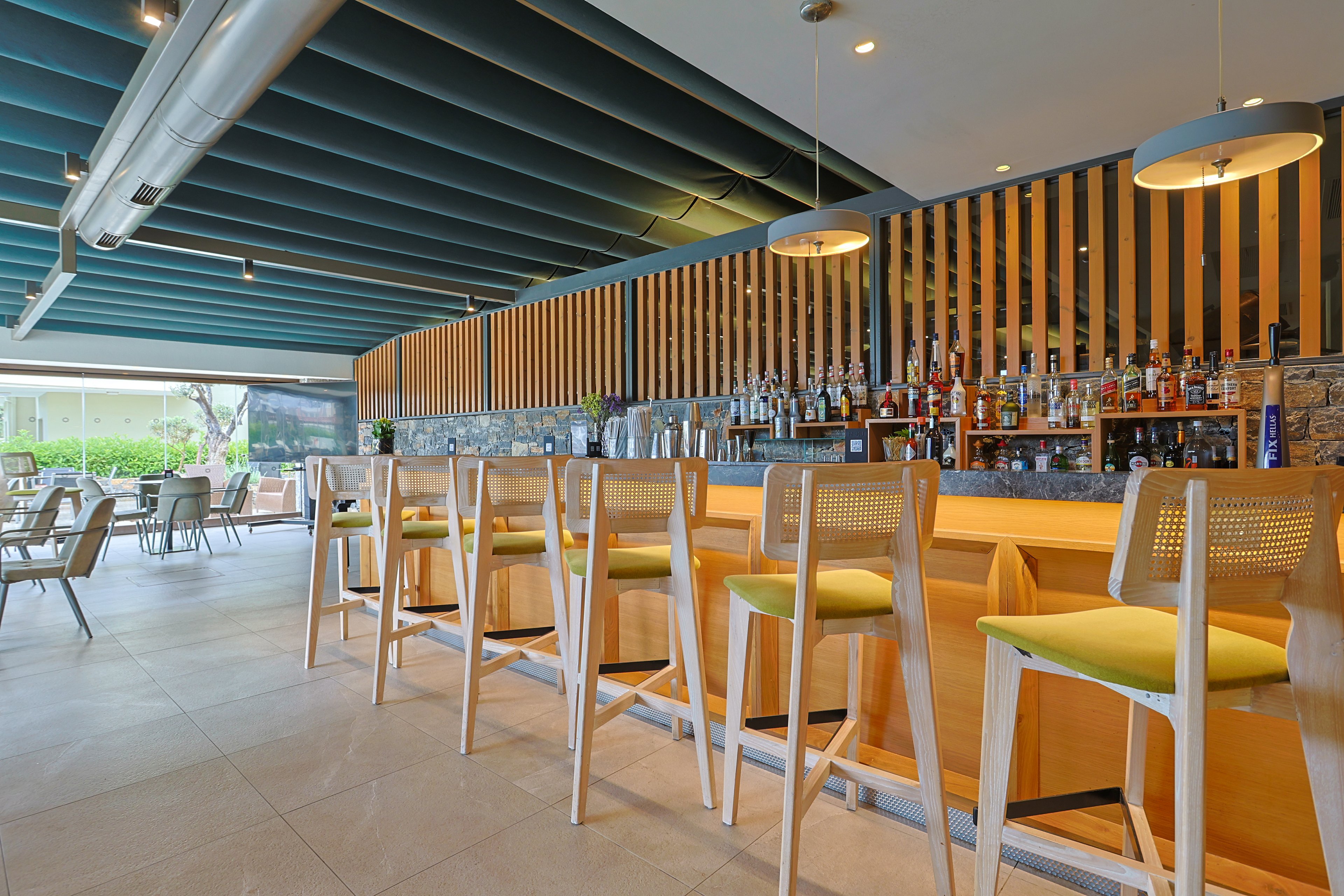 A contemporary bar interior features a long wooden counter lined with light wooden barstools with green cushions. Behind the counter, shelves display an assortment of bottles against a backdrop of vertical wooden slats and stone tiles. Large pendant lights hang from a teal ceiling, illuminating the clean and minimalist design. In the background, an open seating area with tables and chairs looks out toward an outdoor space, suggesting a relaxed resort or hotel setting.