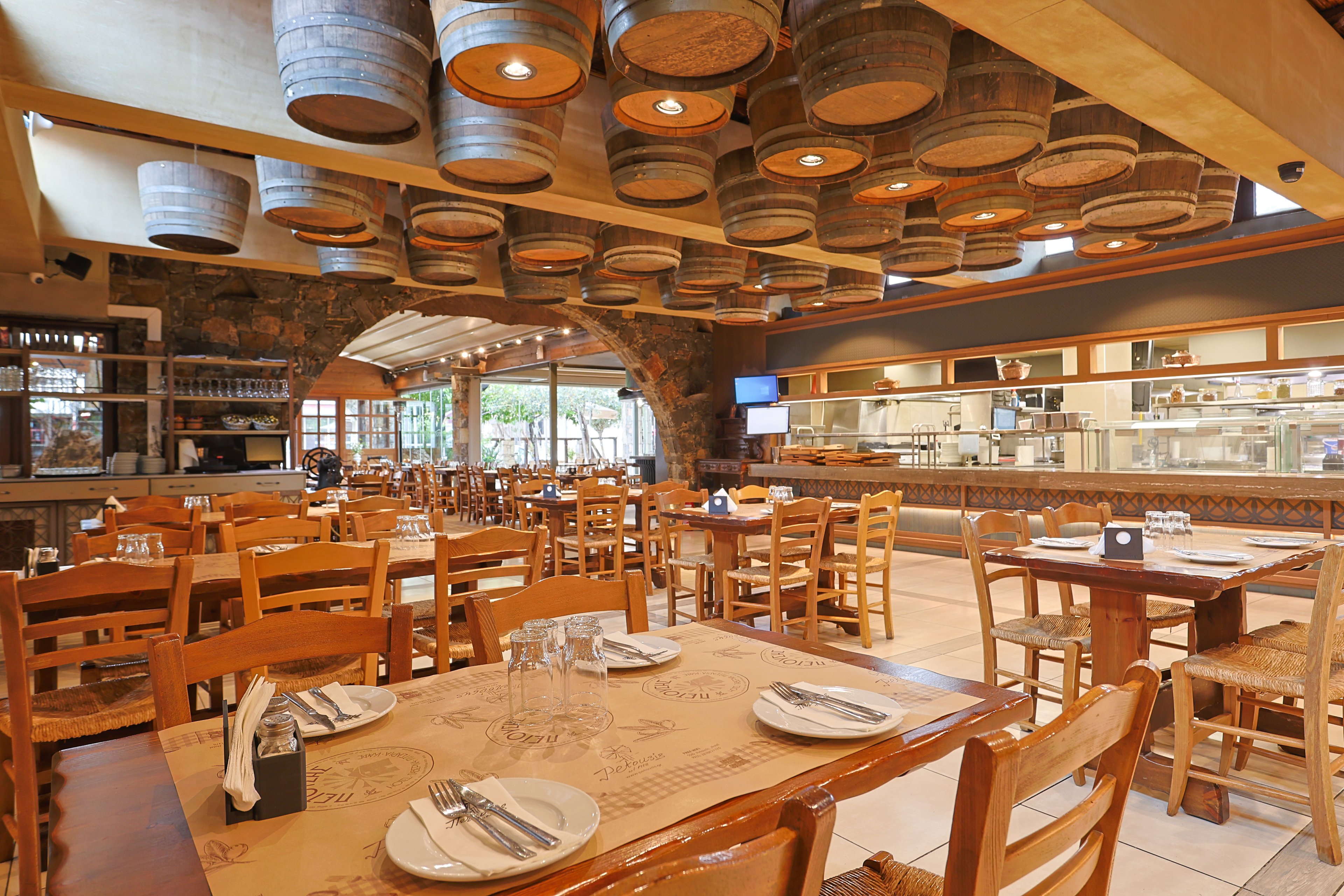 A warm, rustic restaurant dining room features wooden tables and chairs neatly arranged and set with plates, cutlery, and glasses. The ceiling is decorated with numerous wooden barrels, adding a distinctive visual element to the space. Large windows and an arched stone wall open toward an outdoor area, while an open kitchen with stainless steel equipment is visible in the background. The overall atmosphere feels inviting, casual, and ready for guests.