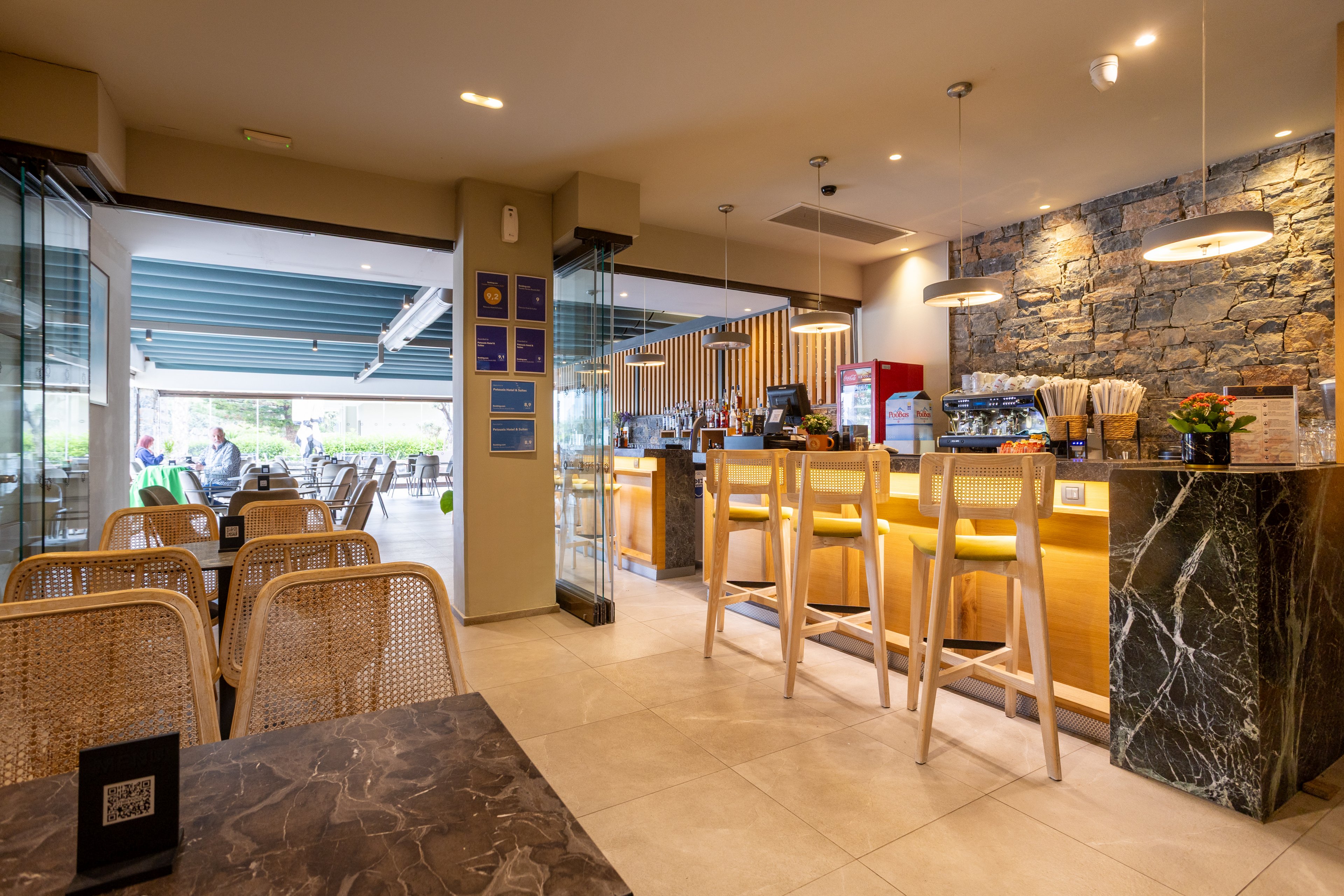 A contemporary cafe interior features a bright bar counter with high wooden stools and warm lighting. The space combines stone and marble textures with clean lines and neutral tones. Large glass doors open to an outdoor seating area, allowing natural light to fill the room. A few patrons are visible in the background, creating a relaxed and welcoming atmosphere.