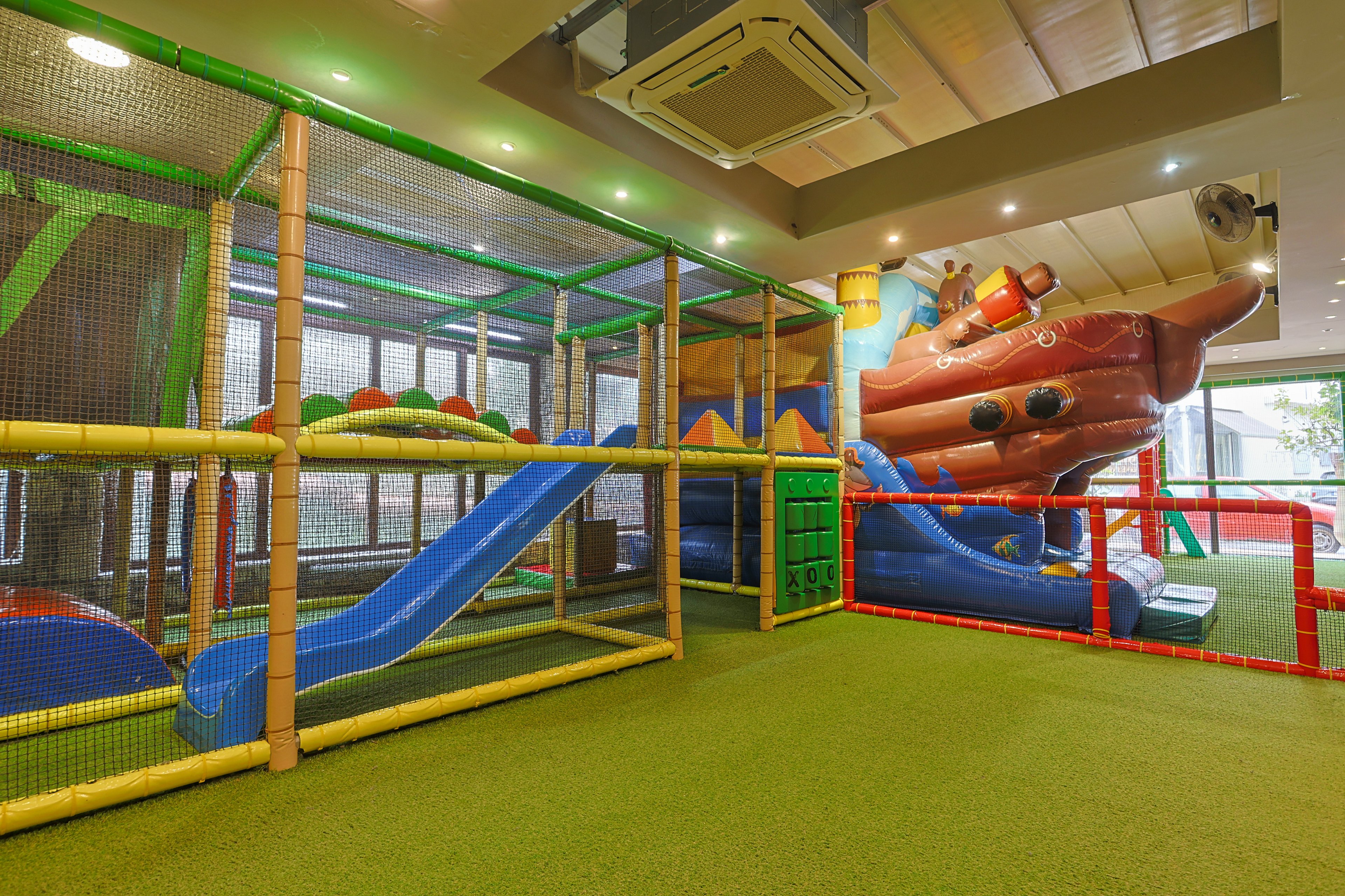 An indoor soft play area features colorful climbing structures and slides. A blue slide and padded platforms are enclosed by safety netting and foam-covered frames. In the background, a large inflatable pirate ship adds a playful focal point. The space is well-lit with artificial lighting and has a soft green floor suitable for children.