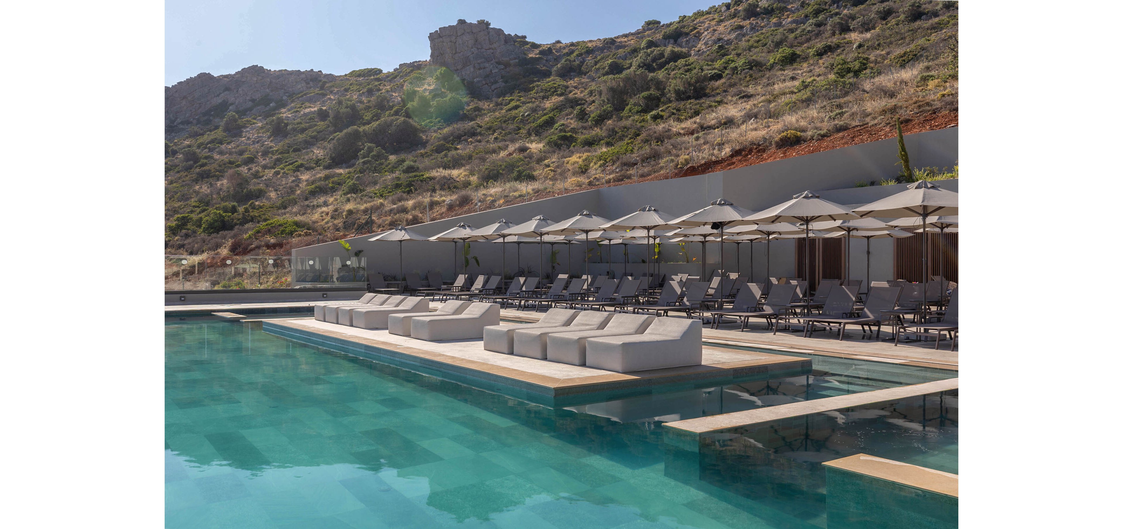A contemporary outdoor swimming pool is surrounded by white cushioned sun loungers and umbrellas. The setting appears to be a luxury resort built at the base of a rocky hillside. Clear turquoise water reflects the minimalist architecture and neutral-toned furniture. The scene is calm and empty, suggesting a peaceful, relaxing atmosphere under bright daylight.