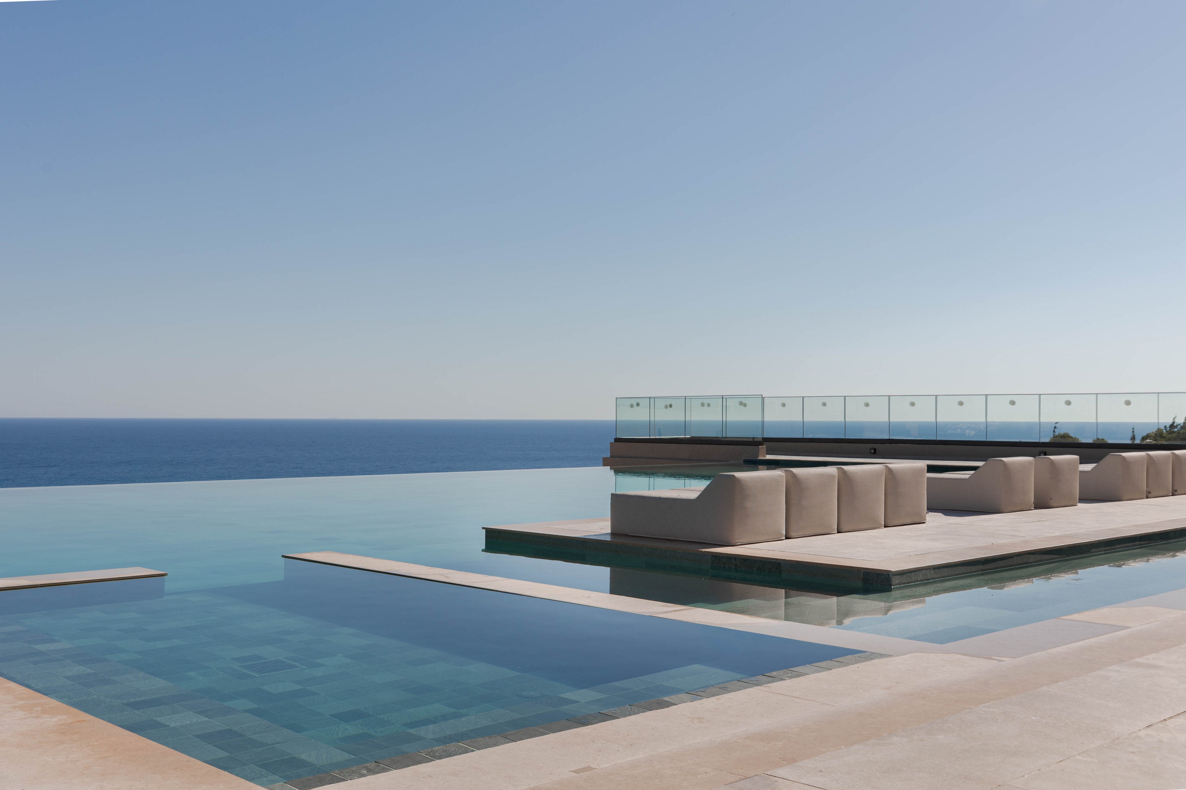 A sleek infinity pool stretches toward a calm blue ocean under a clear sky. Minimalist lounge seating sits on a raised terrace bordered by glass railings. The clean architectural lines and neutral tones create a luxurious, contemporary resort atmosphere. The tranquil water and open horizon emphasize relaxation and coastal serenity.
