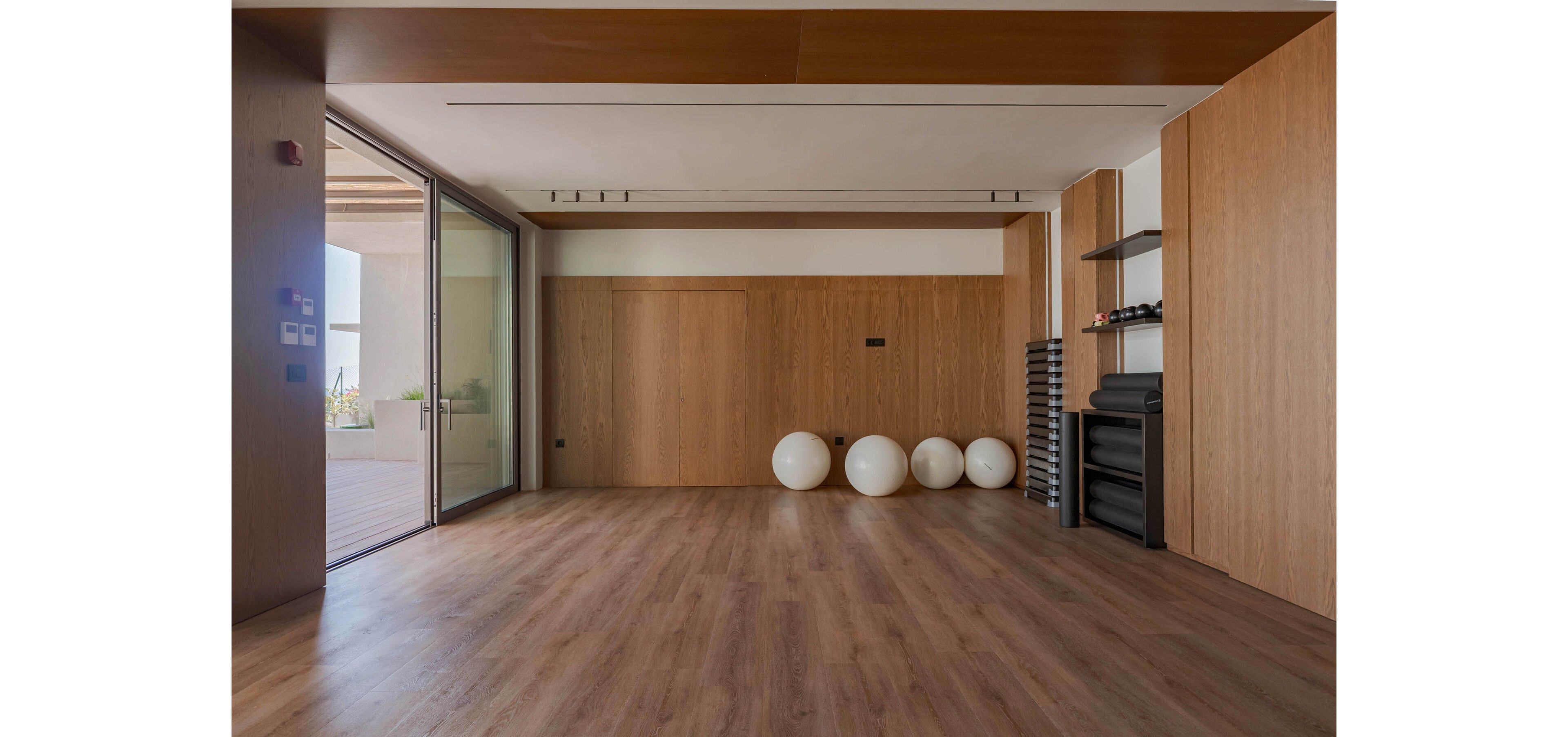 A spacious, minimalist fitness studio features warm wooden floors and wall panels. Several white exercise balls are lined up neatly against the back wall. To the right, stacked mats and shelves with small weights and accessories create an organized workout area. Large sliding glass doors open to an outdoor terrace, filling the room with natural light and a calm, contemporary atmosphere.