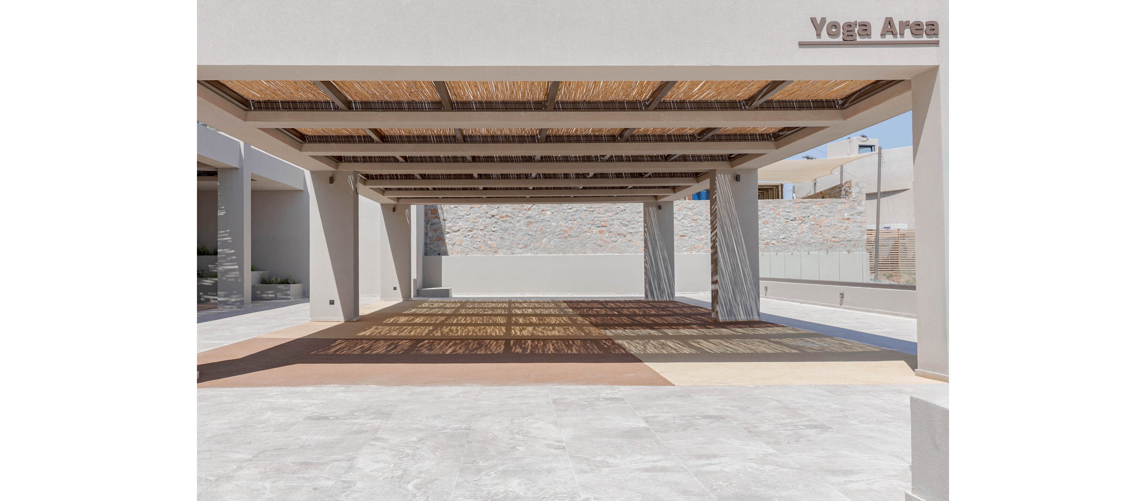 An open-air yoga area is shown under a large shaded pergola structure. The space features clean architectural lines with neutral tones and a smooth floor suitable for exercise. Sunlight filters through the slatted roof, casting geometric shadows across the ground. In the background, a stone wall and glass railings suggest a contemporary resort or wellness facility, and the words "Yoga Area" are visible on the structure.