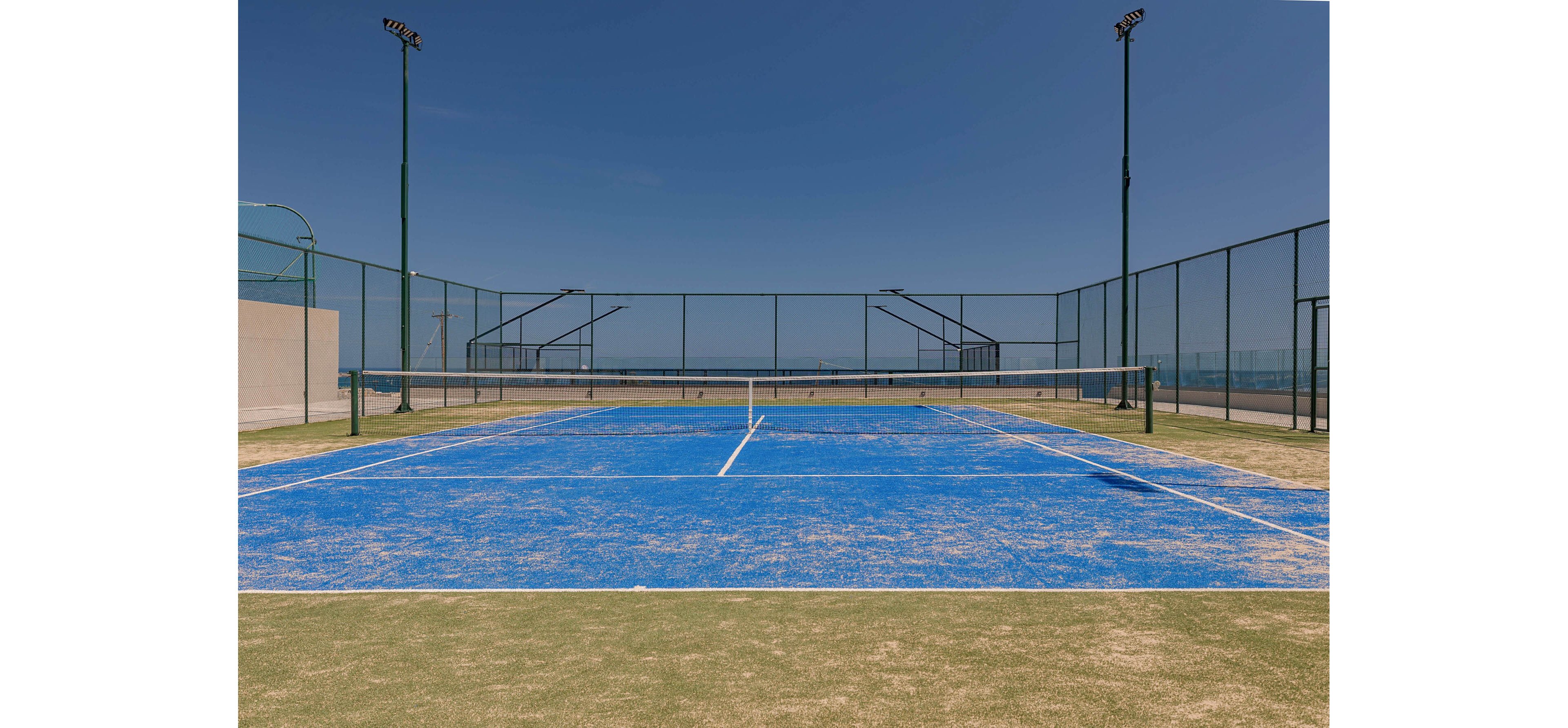 A bright blue tennis court stands empty under a clear blue sky. The court is surrounded by tall chain-link fences and high light poles. Faded lines and worn surface textures suggest frequent use. In the distance, the horizon and subtle landscape are visible beyond the fencing, emphasizing an outdoor sports facility.
