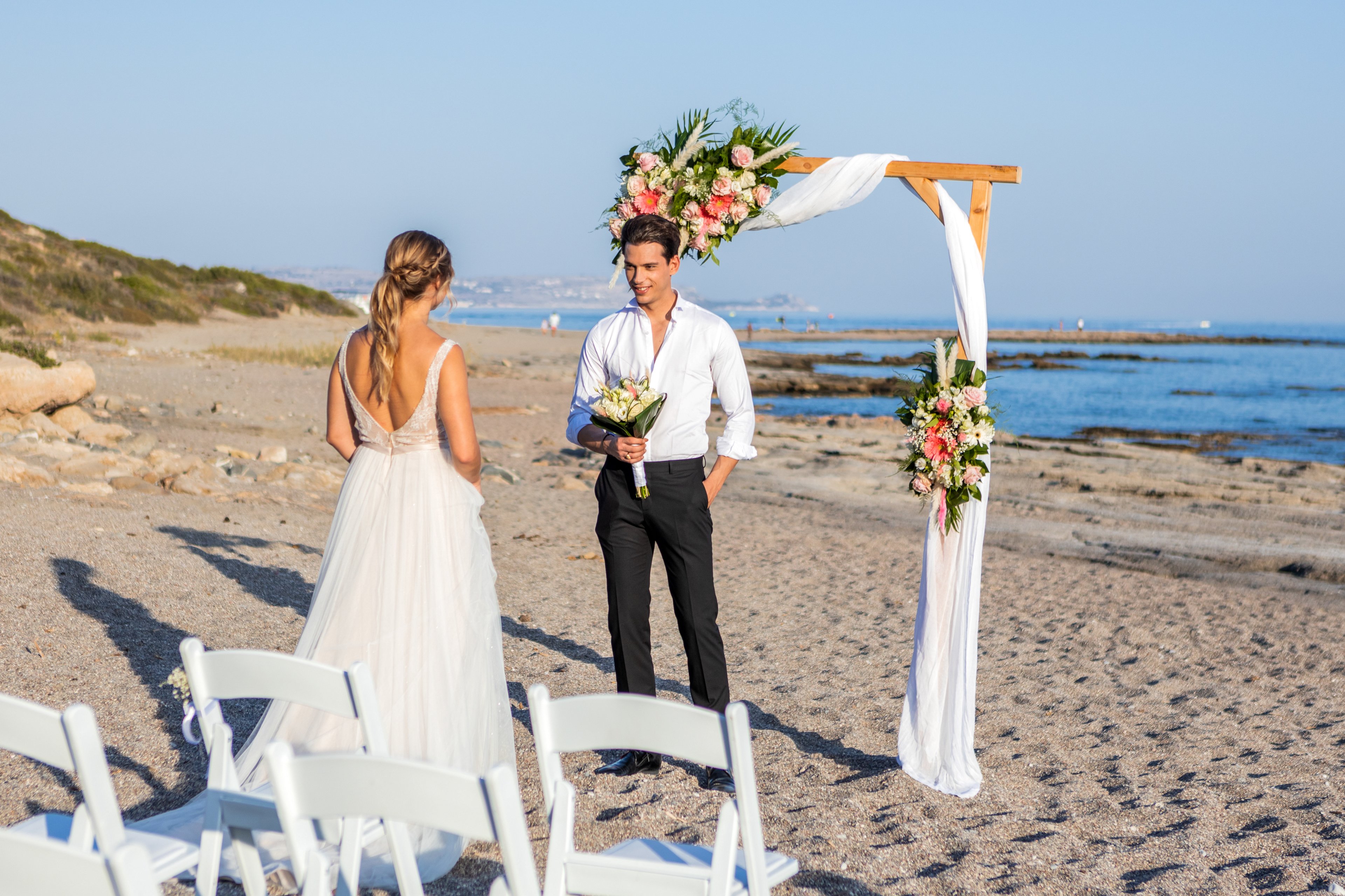A bride in a flowing white gown stands facing a groom on a sandy beach. They are positioned beneath a simple wooden arch decorated with white fabric and pink and white flowers. White folding chairs are arranged in rows, suggesting a small, intimate ceremony. The calm sea and distant coastline create a serene, romantic backdrop under clear blue skies.
