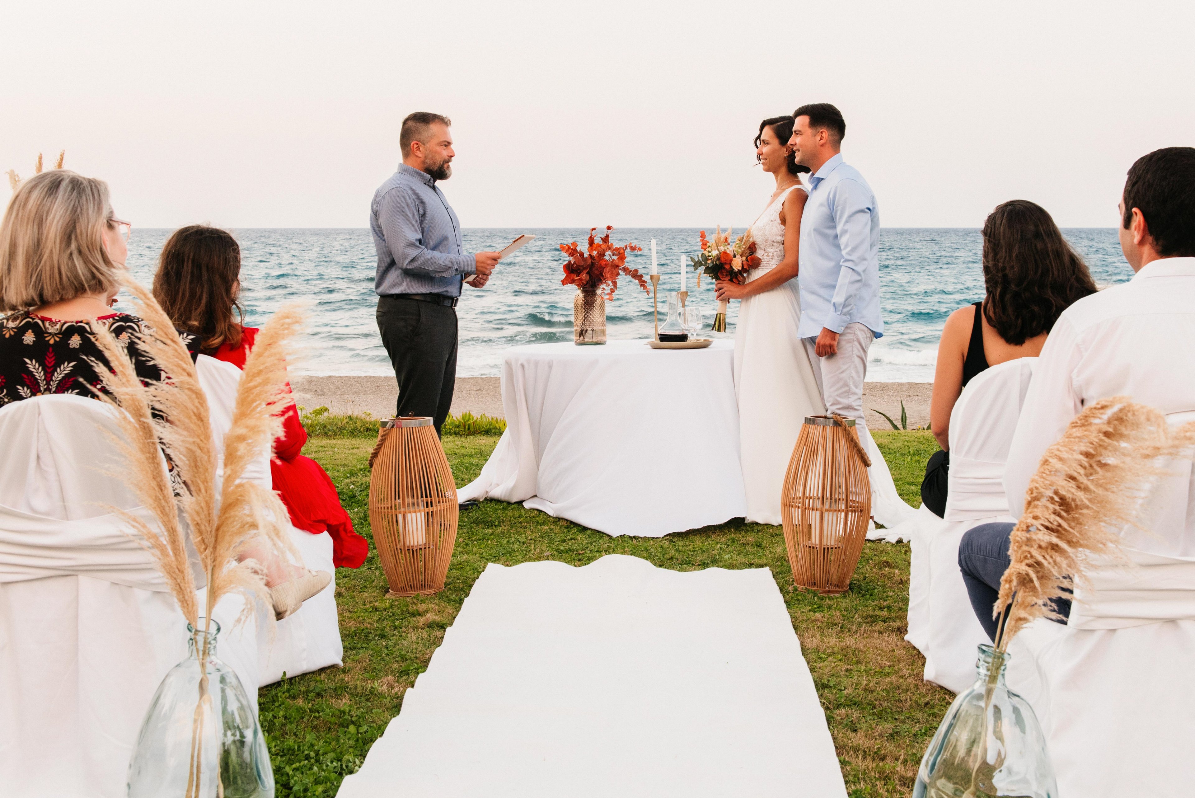 A couple stands together at an altar during an outdoor wedding ceremony by the sea. The officiant faces them while guests sit on white-covered chairs arranged along a white aisle runner. Decorative vases with pampas grass and wooden lanterns frame the aisle, adding a rustic touch. The calm ocean and pastel sky create a serene, romantic backdrop for the event.