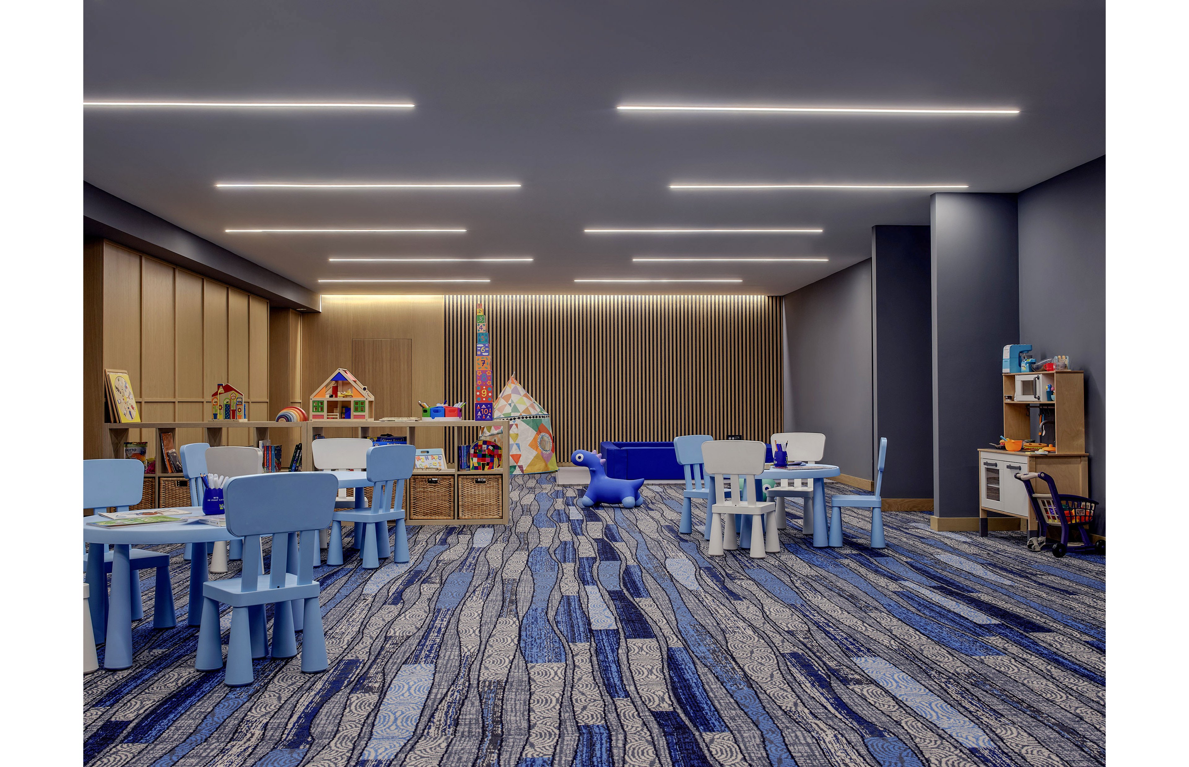 A spacious indoor children's playroom features small blue tables and chairs arranged across a patterned blue carpet. Wooden storage units and shelves hold colorful toys, blocks, and playsets along the walls. Large play structures and a cushioned seating area create a welcoming environment for kids. Recessed ceiling lights provide bright, even illumination, highlighting the clean and modern design of the room.