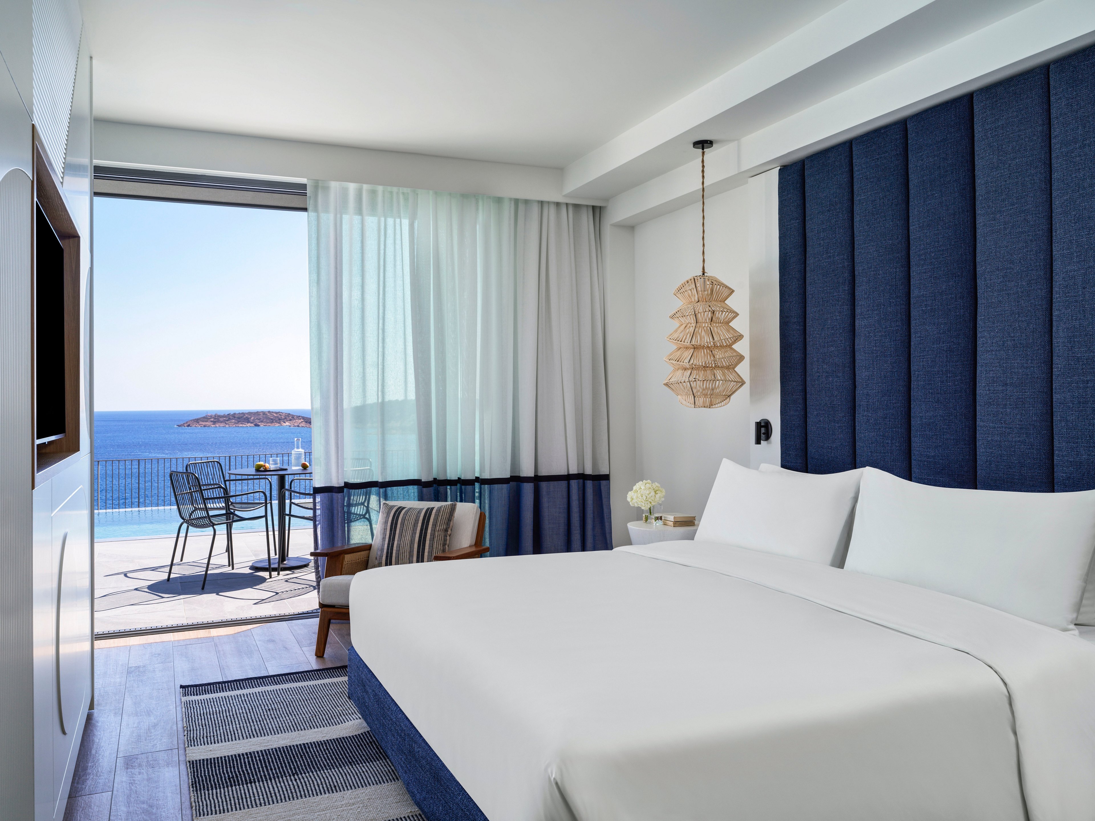 A contemporary hotel room features a large neatly made bed facing a balcony with an expansive ocean view. The interior decor combines white walls with deep blue upholstered panels and a striped rug for a coastal aesthetic. A small seating area with an armchair and side table sits near the sliding glass doors, while a pendant light adds warmth above. Outside, a table and chairs overlook the calm blue sea under a clear sky, creating a serene vacation atmosphere.