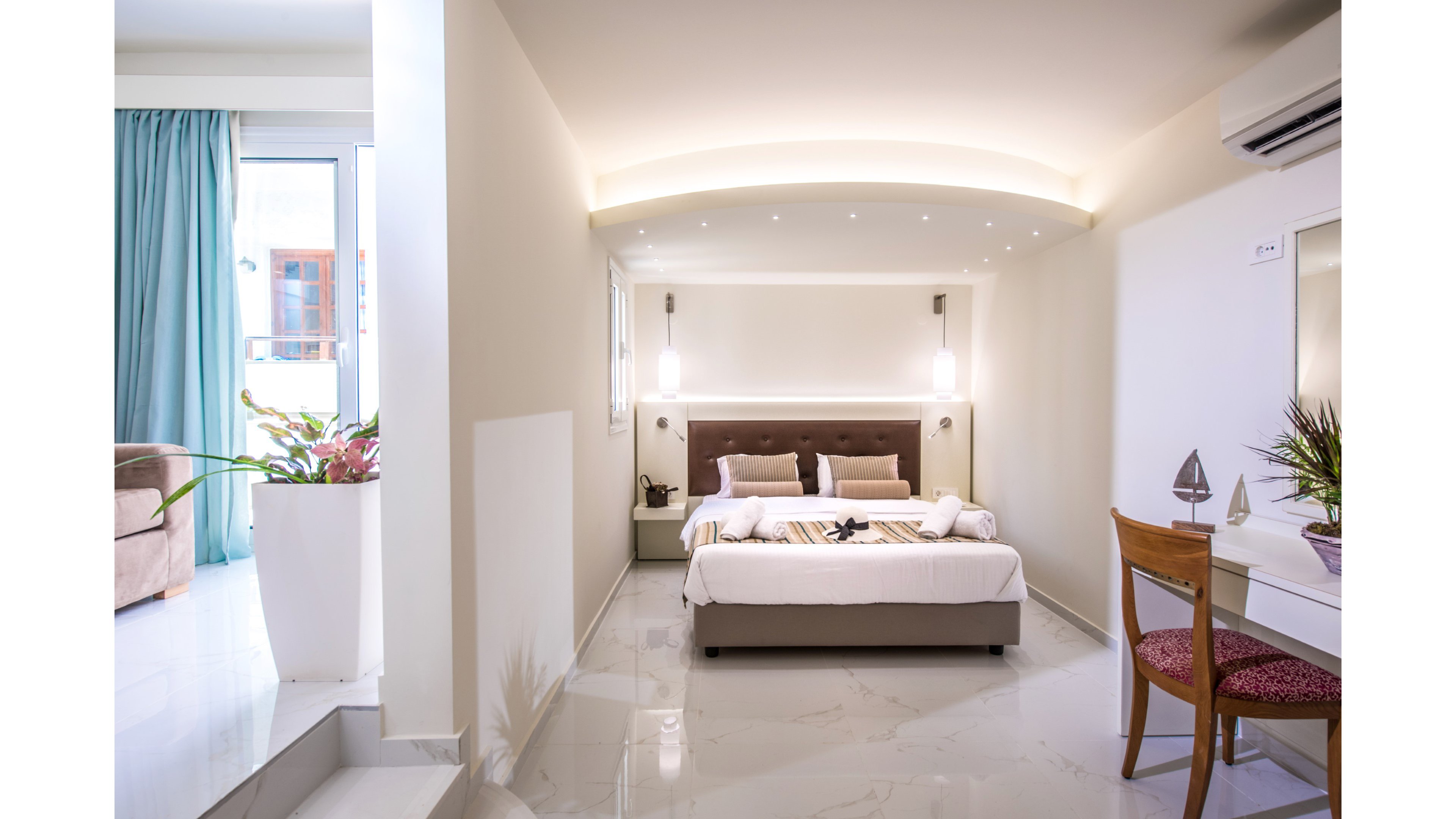 A contemporary hotel-style bedroom features a double bed with plush pillows and folded towels arranged neatly at the foot. Soft pendant lights and recessed ceiling lighting create a warm, inviting atmosphere against neutral walls. A small desk with a chair and decorative items sits to the right, while a large potted plant and blue curtains frame the bright window area. The glossy floor and minimal furnishings emphasize a clean, modern design ideal for relaxation.