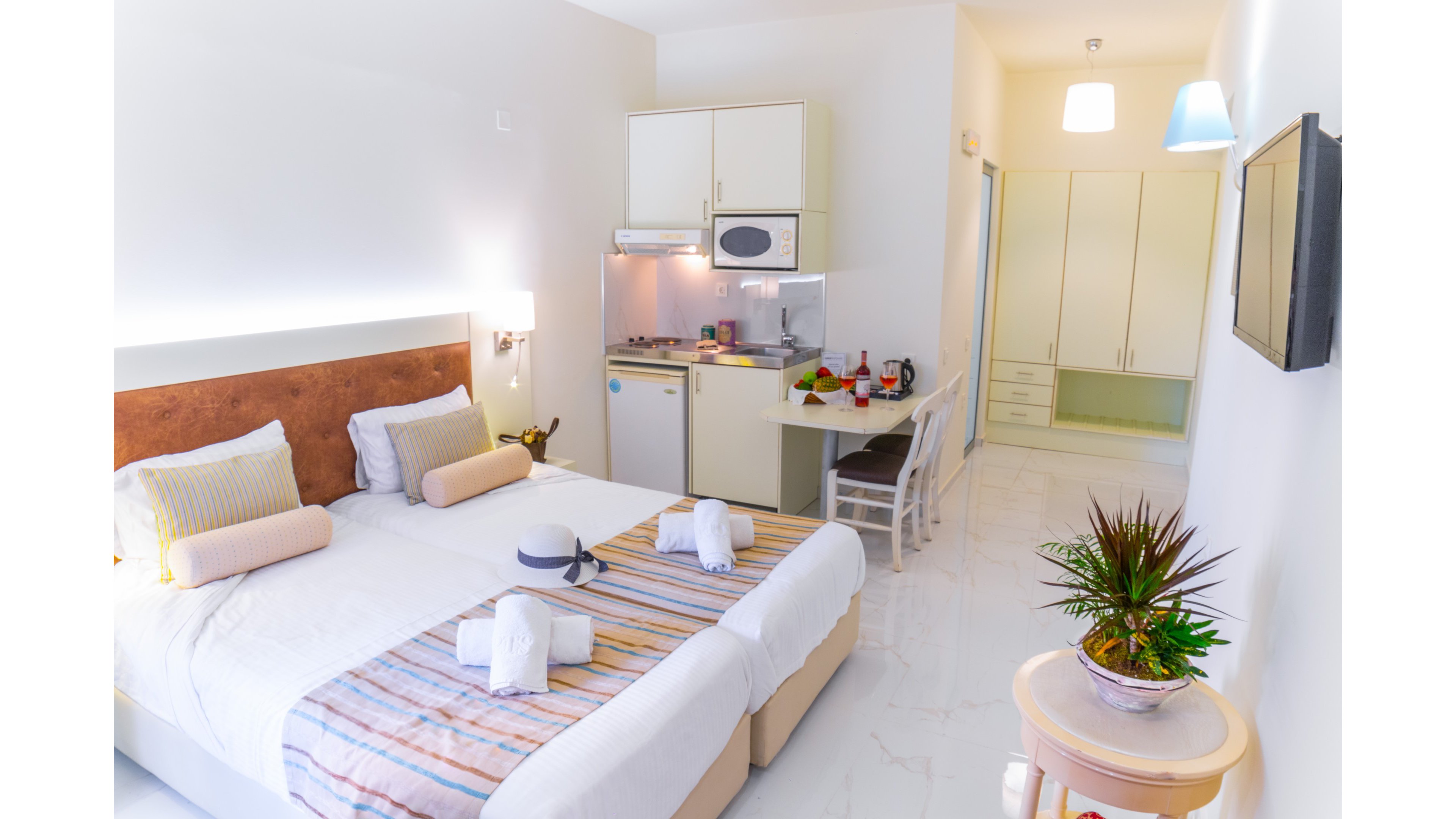 A clean, contemporary hotel room features two twin beds neatly made with striped runners and decorative pillows. A compact kitchenette with cabinets, sink, stovetop, and microwave is set against the wall beside a small desk and chair. The space is brightly lit with white walls, glossy flooring, and soft accent lighting, creating an airy, welcoming atmosphere. A potted plant and folded towels add a touch of comfort and detail to the interior decor.