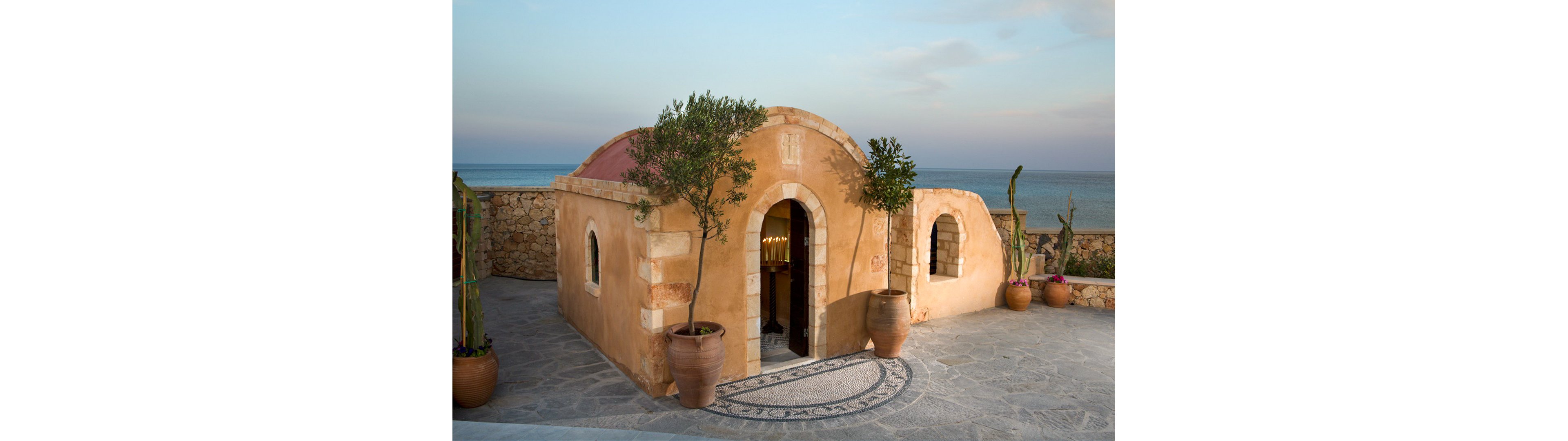 A small stone cottage with a rounded roof stands on a paved terrace by the sea. The building features an arched doorway and small windows framed with light-colored stone. Large terracotta pots with trees and cacti decorate the entrance, and a circular mosaic pattern is set into the ground. In the background, calm blue water and a soft sky create a tranquil coastal atmosphere.