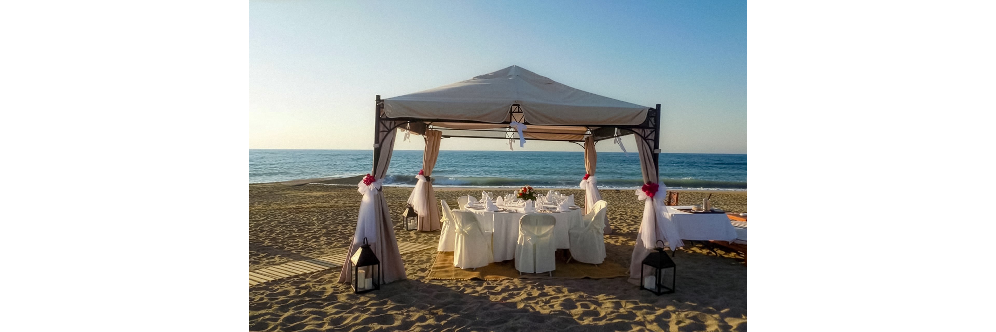 A decorated gazebo stands on a sandy beach, sheltering a round dining table set with white linens and glassware. The calm sea and clear sky create a serene coastal backdrop as the sun nears the horizon. Chairs are draped in white covers, and the gazebo posts are adorned with soft fabric and floral accents. Lanterns rest on the sand near the entrance, enhancing the romantic, intimate atmosphere.