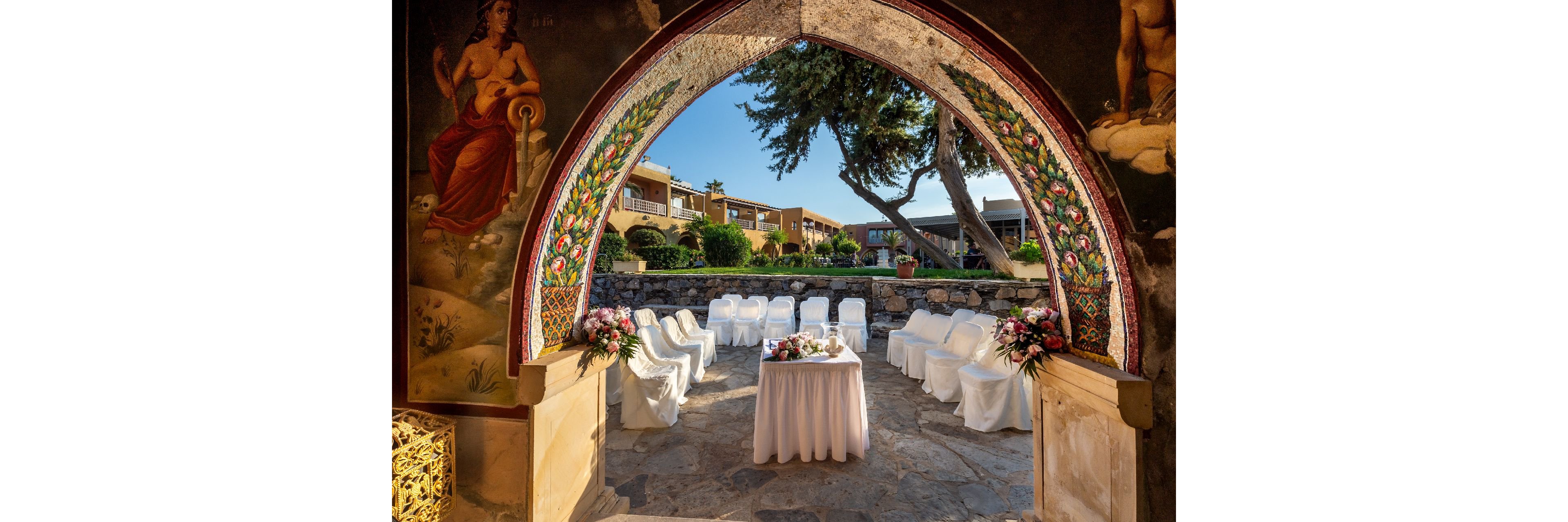 An elegant outdoor wedding ceremony is arranged beneath a decorative stone archway with colorful floral artwork. White-covered chairs form a semi-circle around a central table draped in white cloth and adorned with flowers. The setting is a sunny courtyard with stone flooring, lush greenery, and surrounding buildings in the background. Warm natural light enhances the festive and romantic atmosphere of the scene.