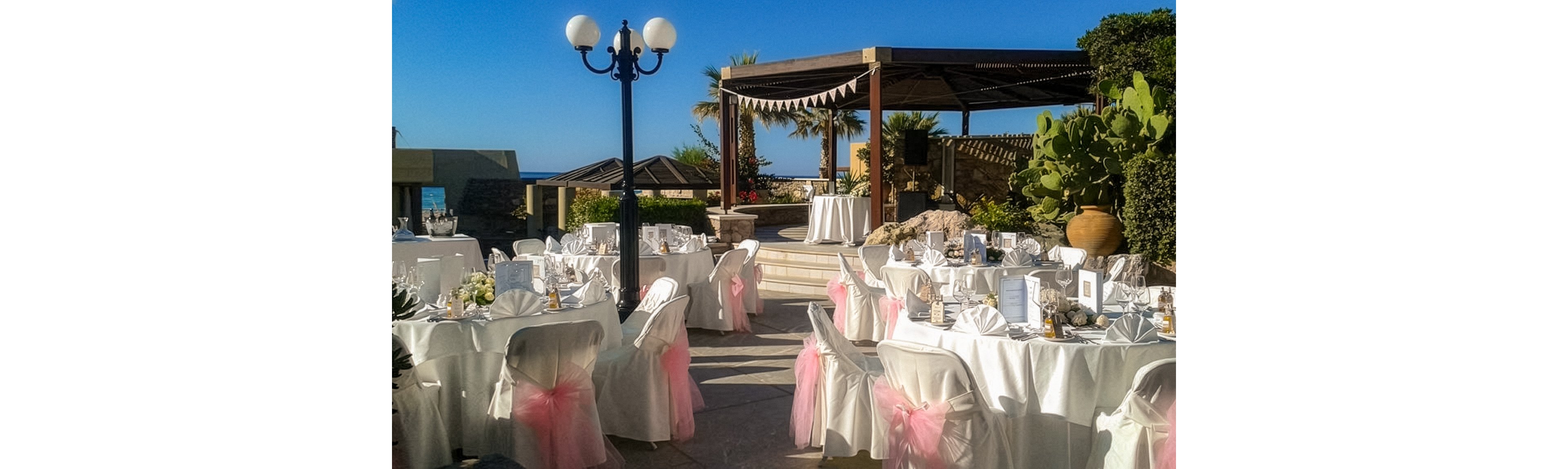 An elegant outdoor wedding reception is set up with round tables covered in white linens and chairs adorned with pink tulle bows. The venue appears to be on a terrace overlooking the sea, with a wooden pergola and bunting in the background. Centerpieces, menus, and small favors decorate each table, creating a festive and romantic atmosphere. Bright daylight and clear blue skies enhance the cheerful, celebratory mood of the event.