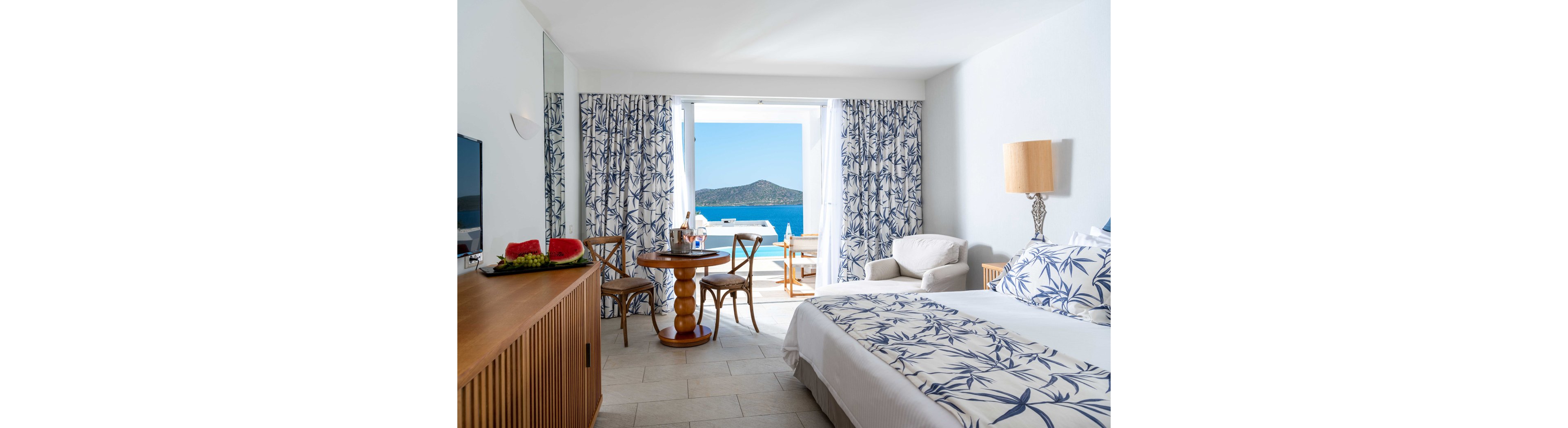 A sunlit hotel bedroom features a neatly made bed with blue and white botanical linens. Matching curtains frame a large open doorway that leads to a balcony overlooking a calm blue sea and distant island. A small wooden table with chairs is set near the balcony, holding drinks and a wine bottle, while an armchair and side lamp complete the cozy seating area. A sideboard with a television and a plate of fresh fruit adds comfort and a welcoming touch to the serene coastal setting.