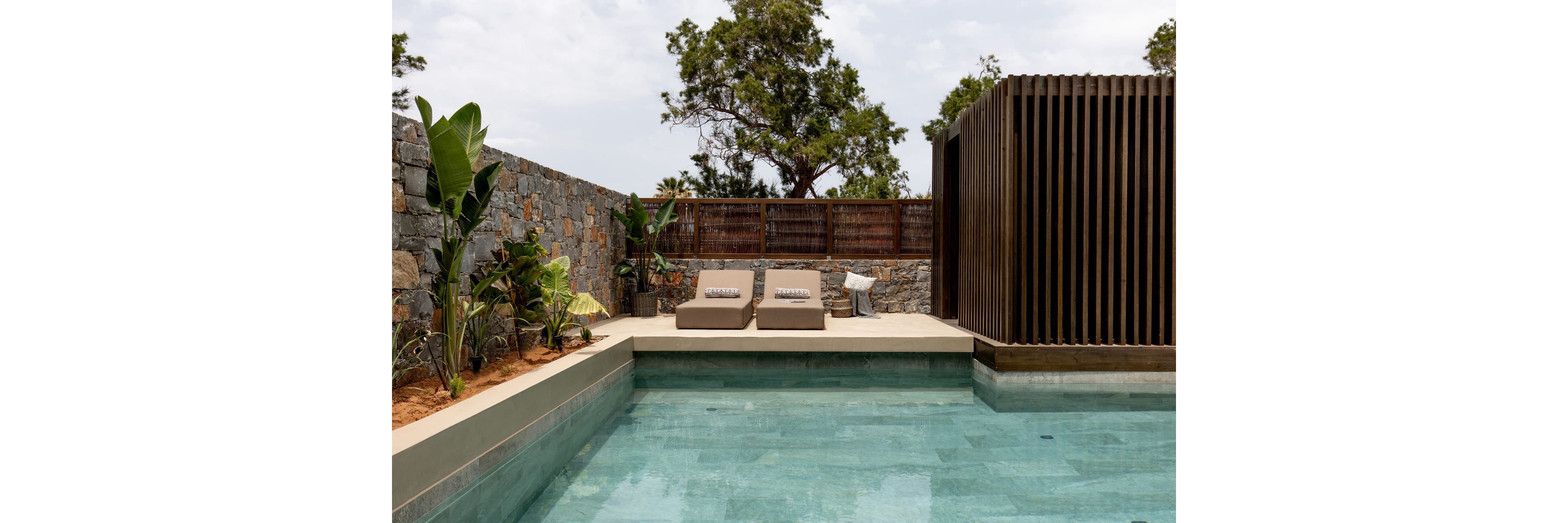 A contemporary outdoor pool is framed by smooth concrete edges and clear blue water. Two cushioned sun loungers sit on the terrace, facing the pool for relaxation. A stone wall, wooden fence, and vertical slatted structure create a secluded, modern courtyard. Lush green plants soften the hardscape, while trees rise above the enclosure under a bright sky.