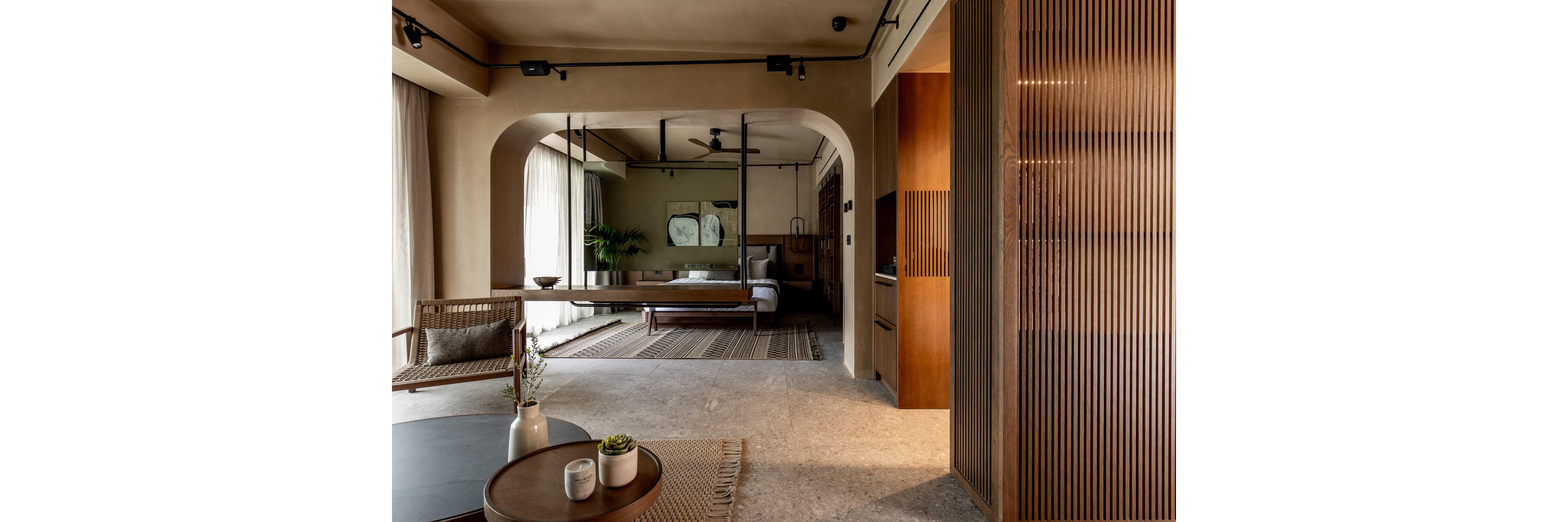 A contemporary open-plan bedroom suite features a bed centered between large windows with sheer curtains. Warm wooden slats, built-in cabinetry, and minimalist furniture create a calm, sophisticated atmosphere. Neutral tones, textured rugs, and indoor plants add softness and natural elements to the space. Soft daylight filters through the curtains, highlighting the clean lines and modern design.