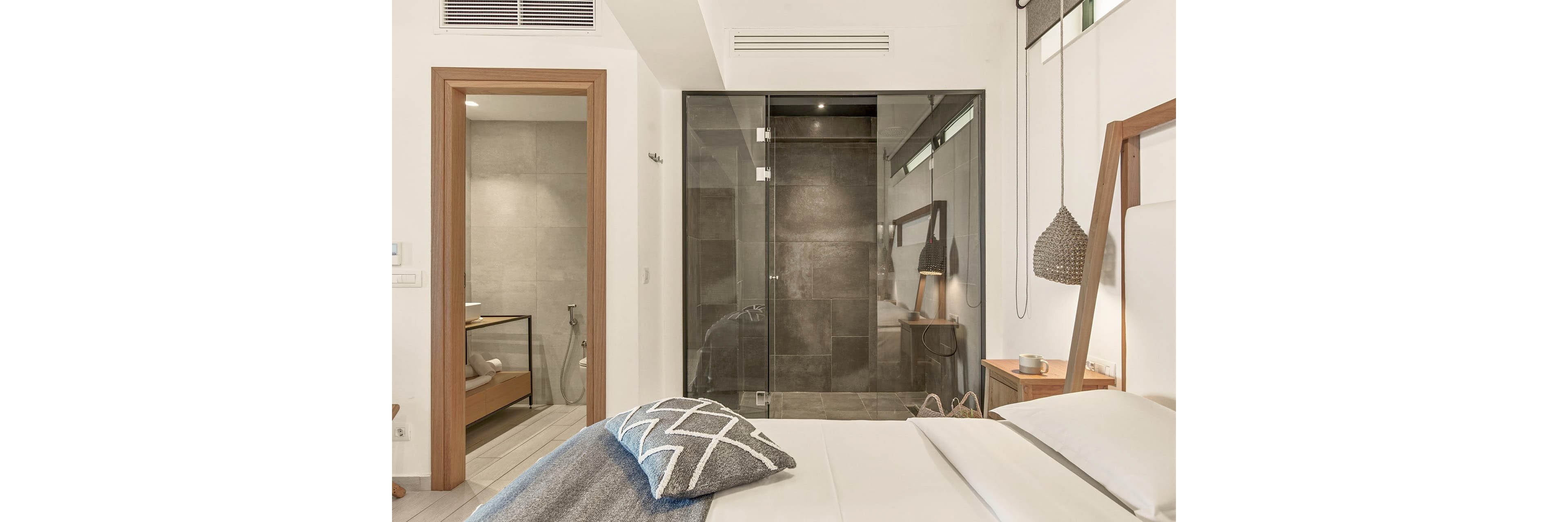 Contemporary bedroom interior features a neatly made bed with white linens and a patterned throw pillow. A glass-enclosed shower with dark tiles is visible behind the bed, alongside a separate bathroom area with light tiles and a handheld shower. Warm wooden accents, a bedside table with accessories, and soft neutral tones create a cozy, minimalist atmosphere. The space is well-lit, highlighting clean lines and modern fixtures.