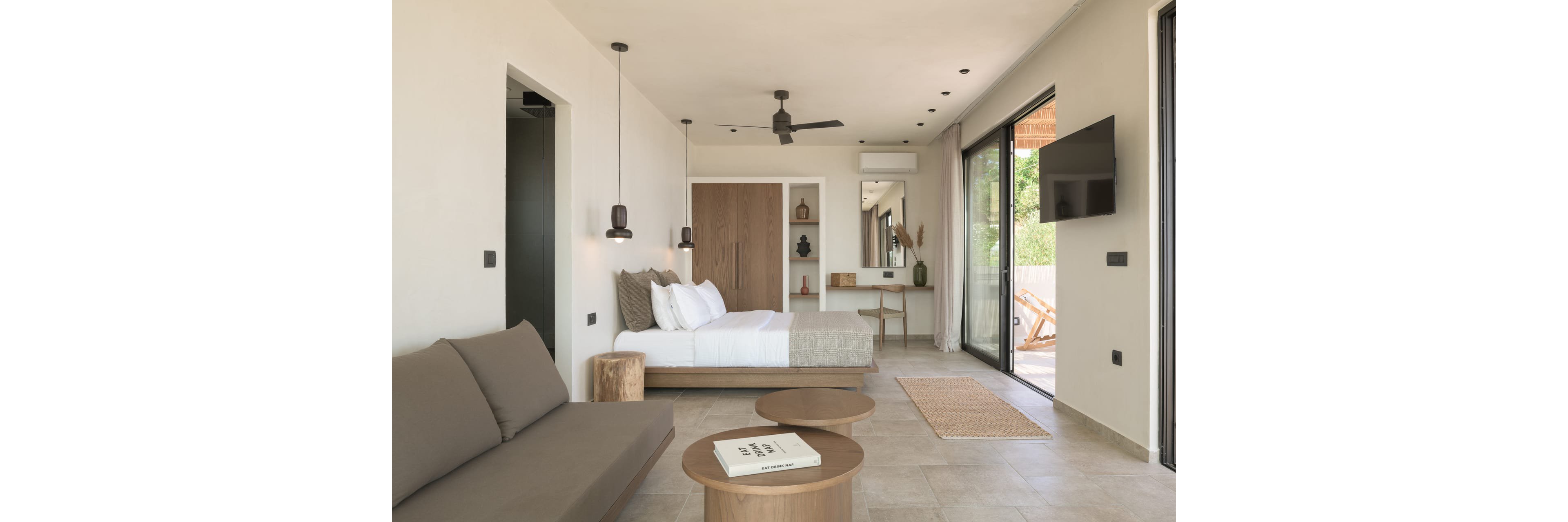 A contemporary hotel room features a neatly made bed, a sofa, and round wooden coffee tables in a neutral-toned interior. Large sliding glass doors open to a terrace with outdoor seating, allowing natural light to fill the space. The room includes a wall-mounted television, ceiling fan, pendant lights, and a small desk area with decorative objects. Soft beige and brown hues create a calm, minimalist atmosphere suitable for relaxation or vacation stays.