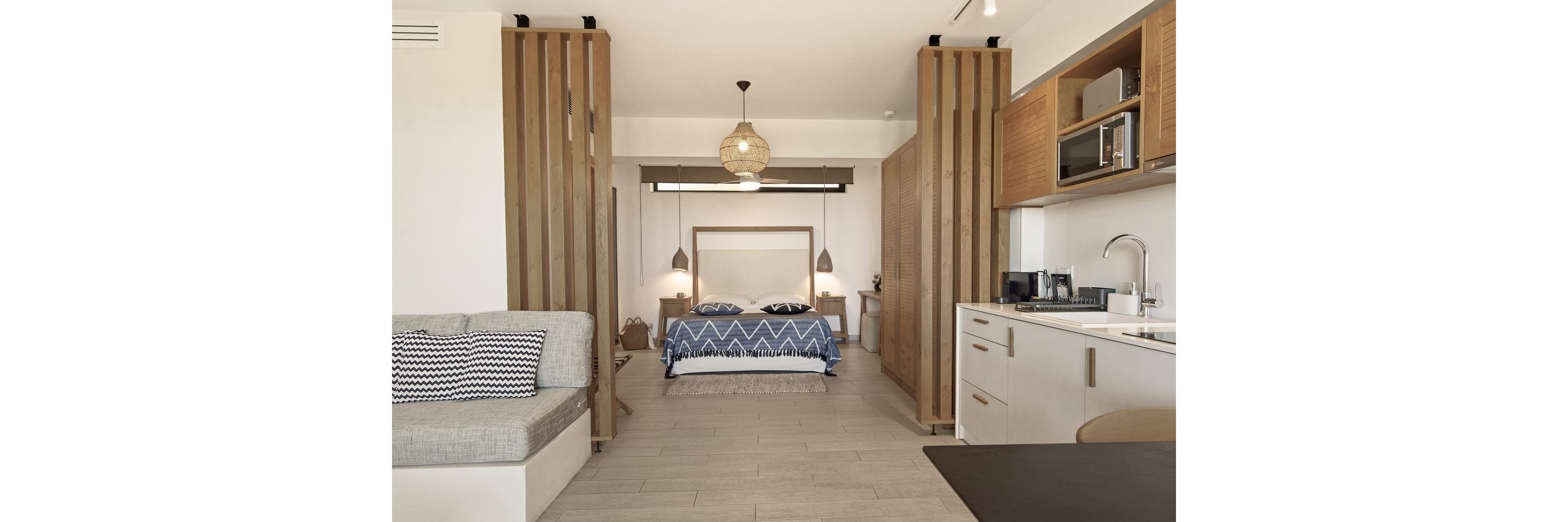 Contemporary studio interior features a central bed with a blue patterned blanket and matching pillows. Wooden slatted partitions subtly separate the sleeping area from the living space and compact kitchen. Neutral tones, light wood flooring, and minimalist furniture create a calm, airy atmosphere. Pendant lights, woven lamp shades, and geometric textiles add stylish decorative accents.