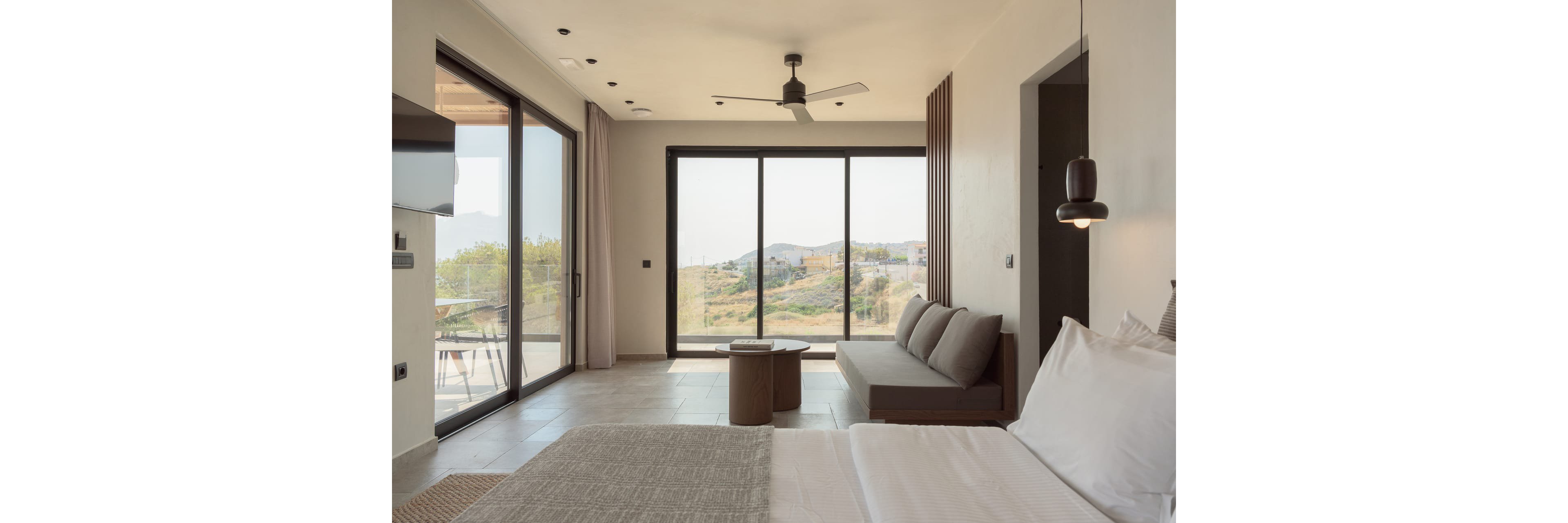 A contemporary hotel room features a neatly made bed and a minimalist sofa facing large floor-to-ceiling windows. The space opens to a balcony with outdoor seating and a scenic view of hills and greenery. Neutral tones, clean lines, and soft natural light create a calm, airy atmosphere. Subtle decorative elements and modern fixtures emphasize a stylish, comfortable retreat.