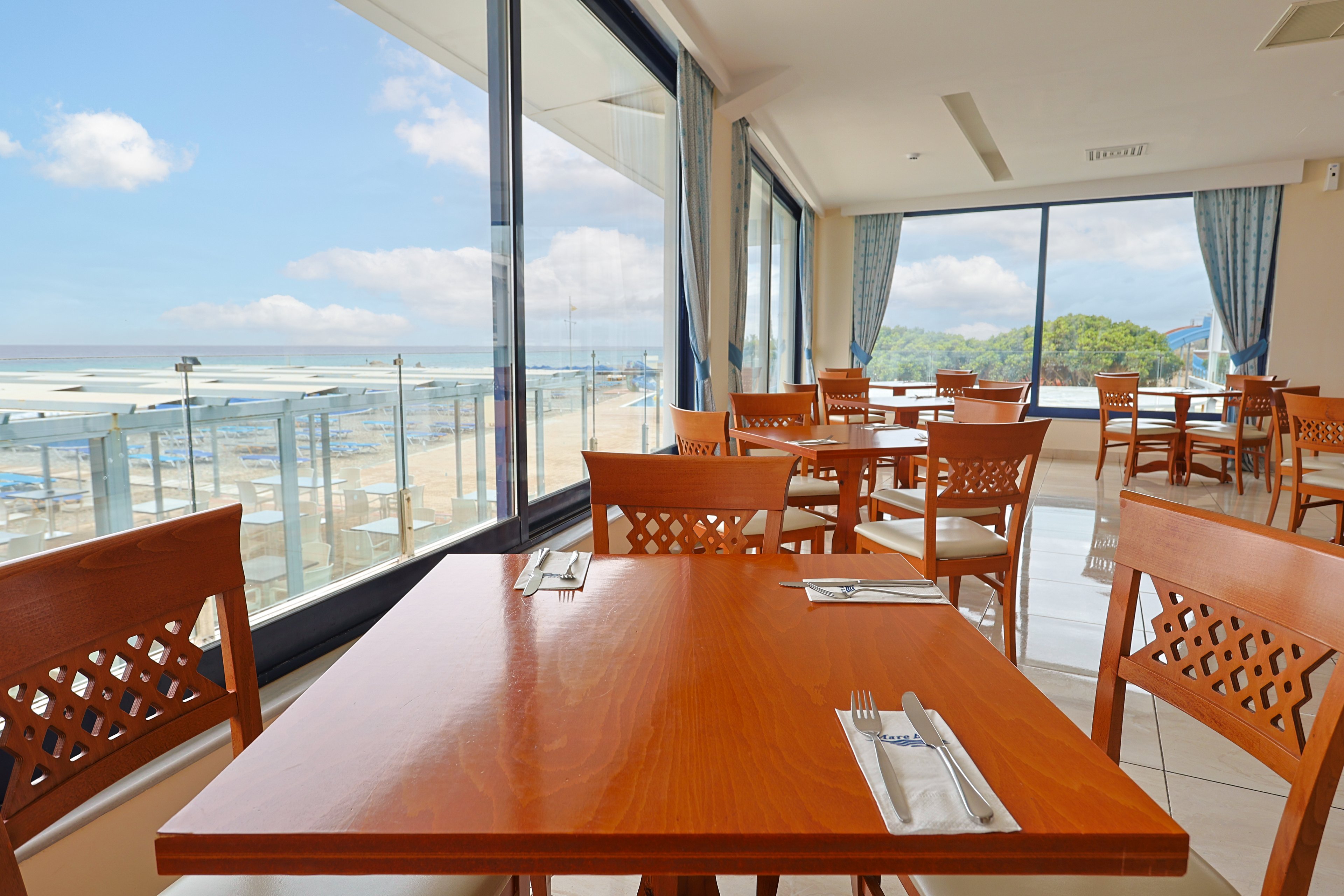 A modern seaside restaurant features wooden tables neatly set with cutlery and napkins. Large floor-to-ceiling windows reveal a bright oceanfront view with sandy beach and blue sky. Sunlight fills the airy dining room, highlighting the clean, minimalist decor. The scene feels calm and inviting, ideal for relaxed coastal dining.