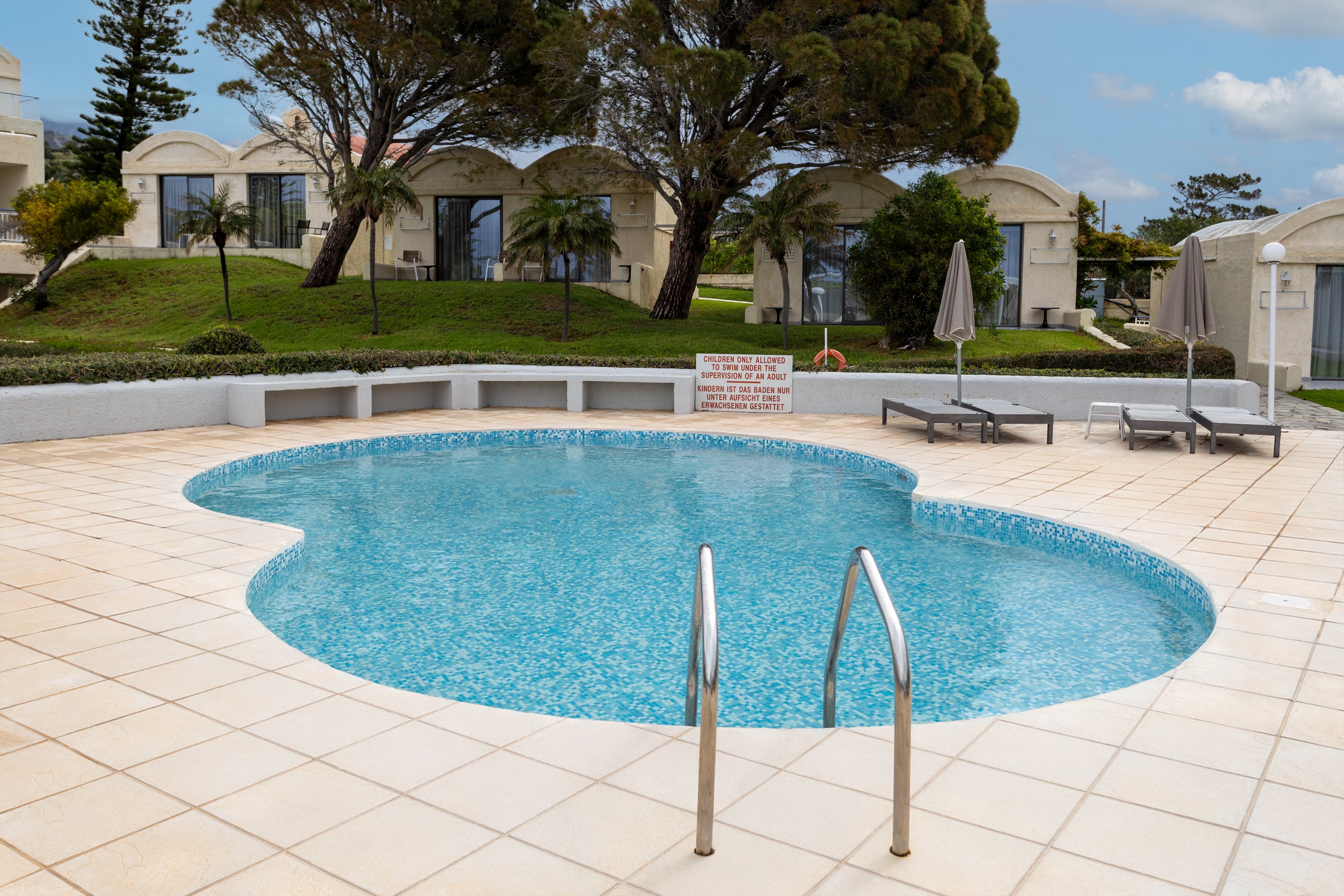 A small outdoor swimming pool sits in the center of a tiled patio at a resort or vacation property. Metal handrails lead into the clear blue water, and a curved white bench surrounds part of the pool. Two sun loungers and closed umbrellas are arranged nearby, suggesting a quiet relaxation area. In the background, landscaped grass, trees, and low white buildings complete the tranquil setting. A sign with safety information is mounted on the far side of the pool.