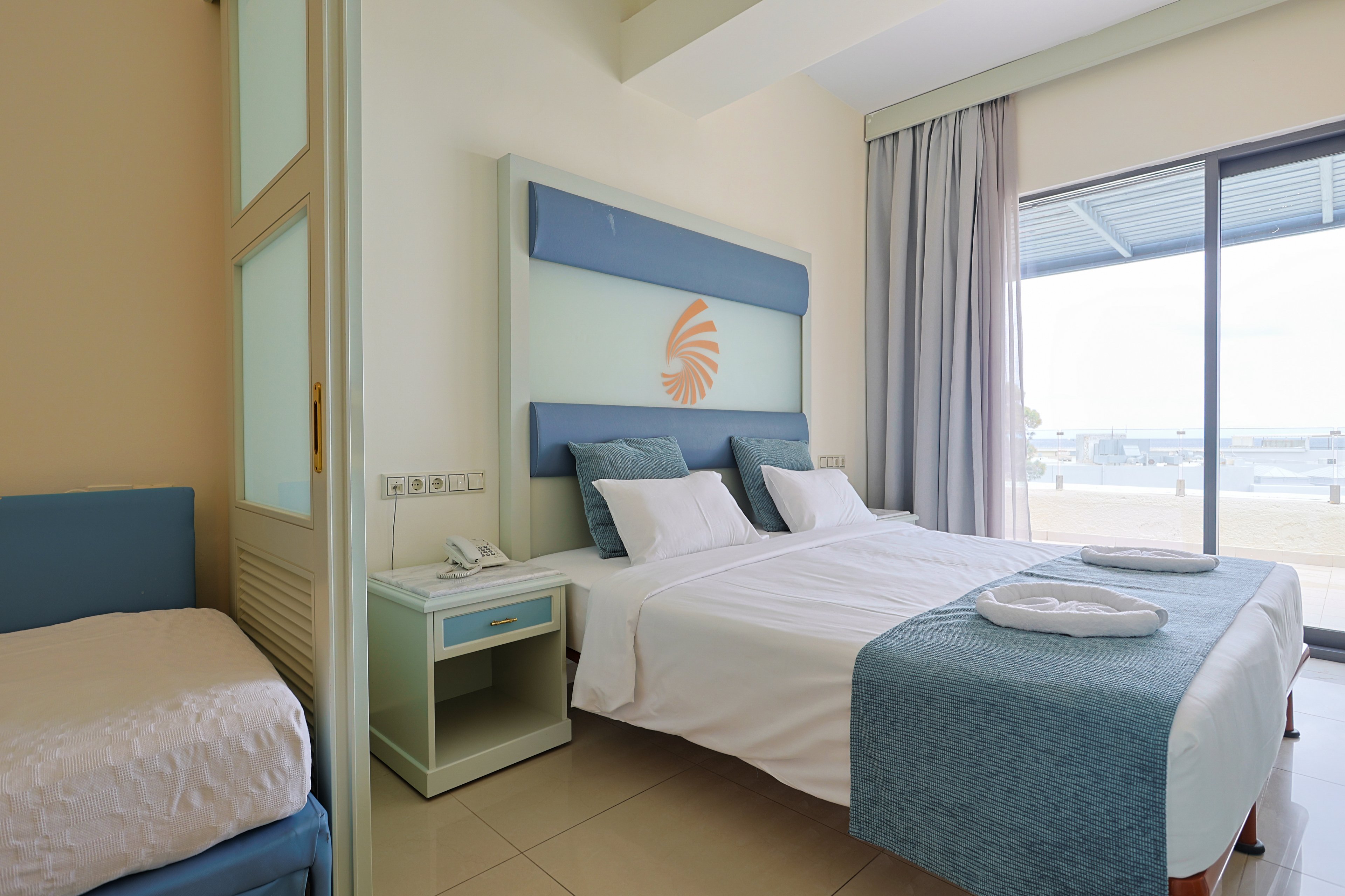 A modern hotel room features a neatly made double bed with white linens and light blue accents. A small bedside table with a telephone sits beside the bed, and folded towels are placed on the foot of the bed. To the left, an additional single bed is partially visible, suggesting accommodation for extra guests. Large sliding glass doors open to a balcony with views of a waterfront or marina area, filling the room with natural light.