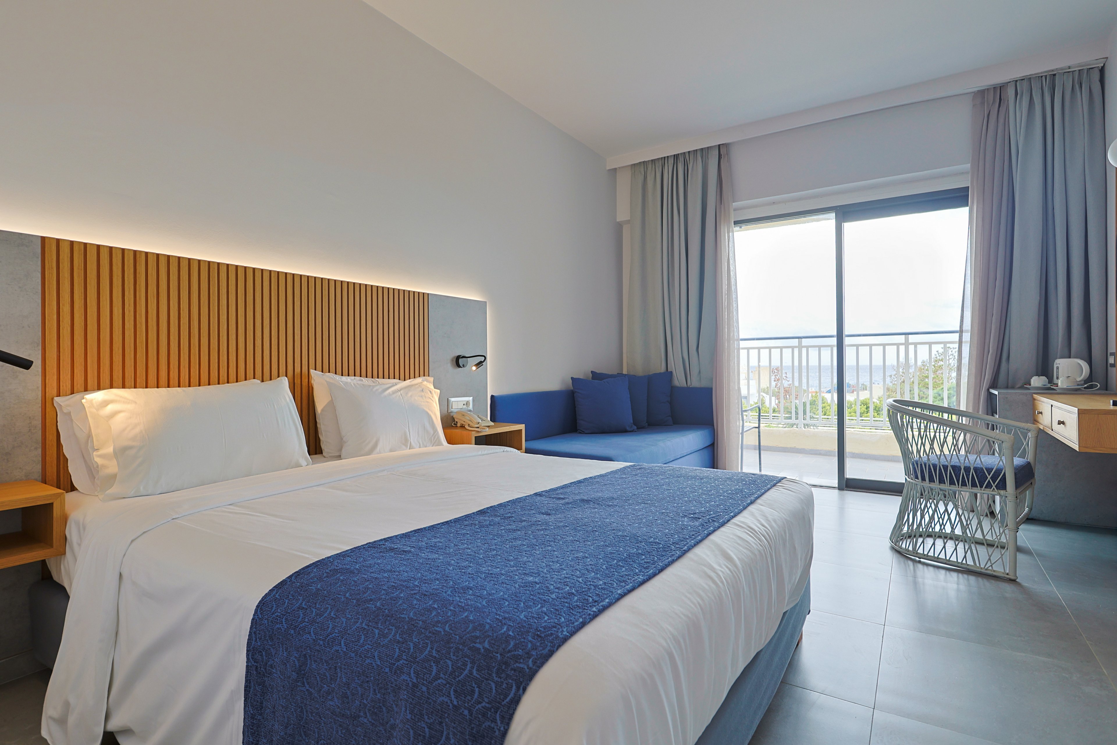 A contemporary hotel room features a neatly made double bed with white linens and a blue bed runner. The space includes a wooden headboard, a blue sofa, and a light-colored tiled floor. A desk with a chair sits near a large sliding glass door that opens to a balcony with a railing and outdoor view. Soft natural light and neutral tones create a calm, inviting atmosphere.