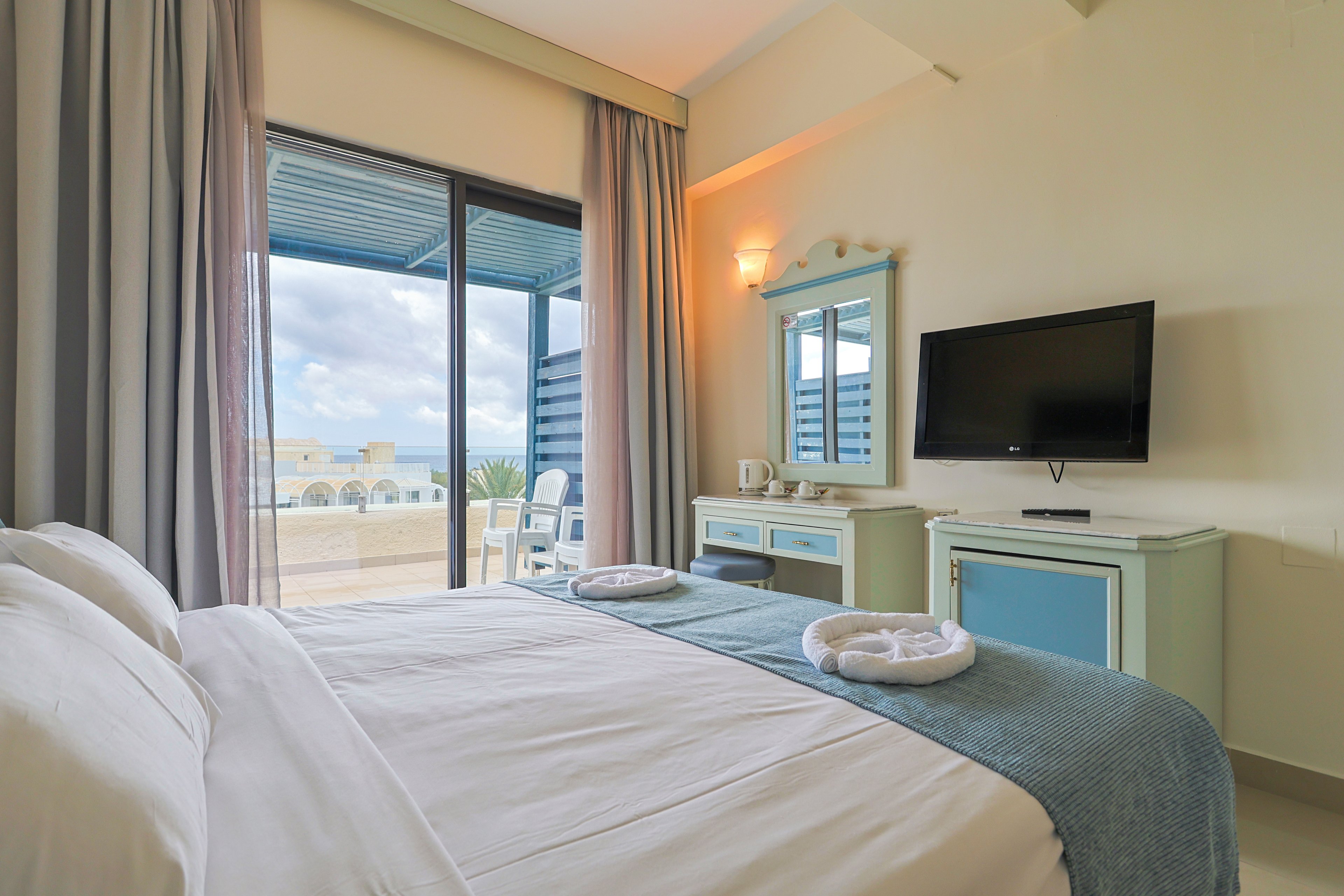 A bright hotel bedroom features a neatly made double bed with folded towels at the foot. The room includes a wall-mounted flat-screen television, a small desk with a mirror, and a tea and coffee station. Large sliding glass doors open onto a terrace with outdoor seating and a view of the cloudy sky and distant buildings. Soft neutral walls and blue accents create a calm, coastal-inspired atmosphere.