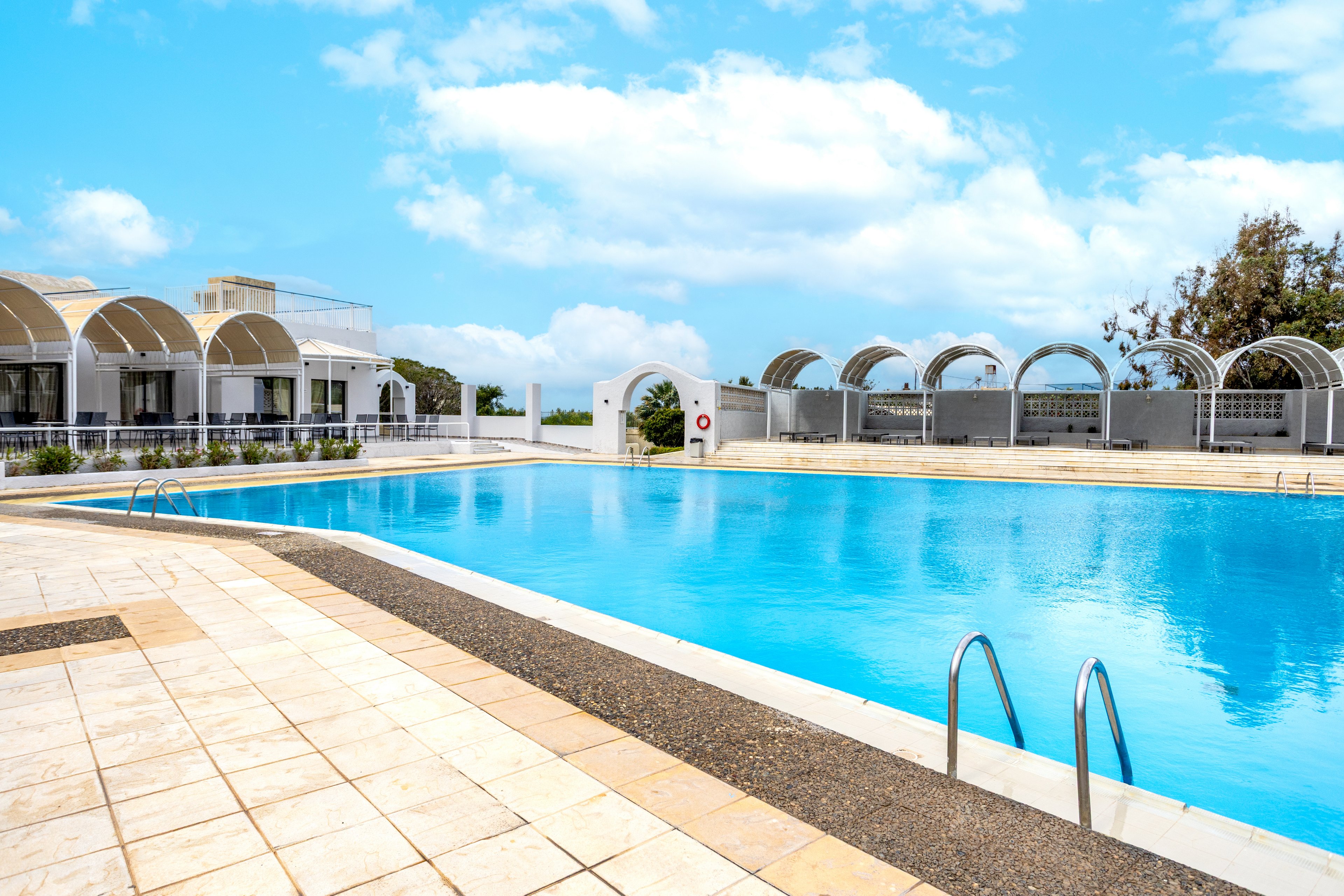 A large outdoor swimming pool dominates the foreground under a bright blue sky with scattered clouds. Modern white arched structures and a low building line the far side of the pool, suggesting a resort or recreational facility. Stainless steel ladders descend into the clear blue water, and the surrounding deck is paved with light-colored tiles. The scene feels calm and inviting, with no people present and strong daylight illuminating the area.