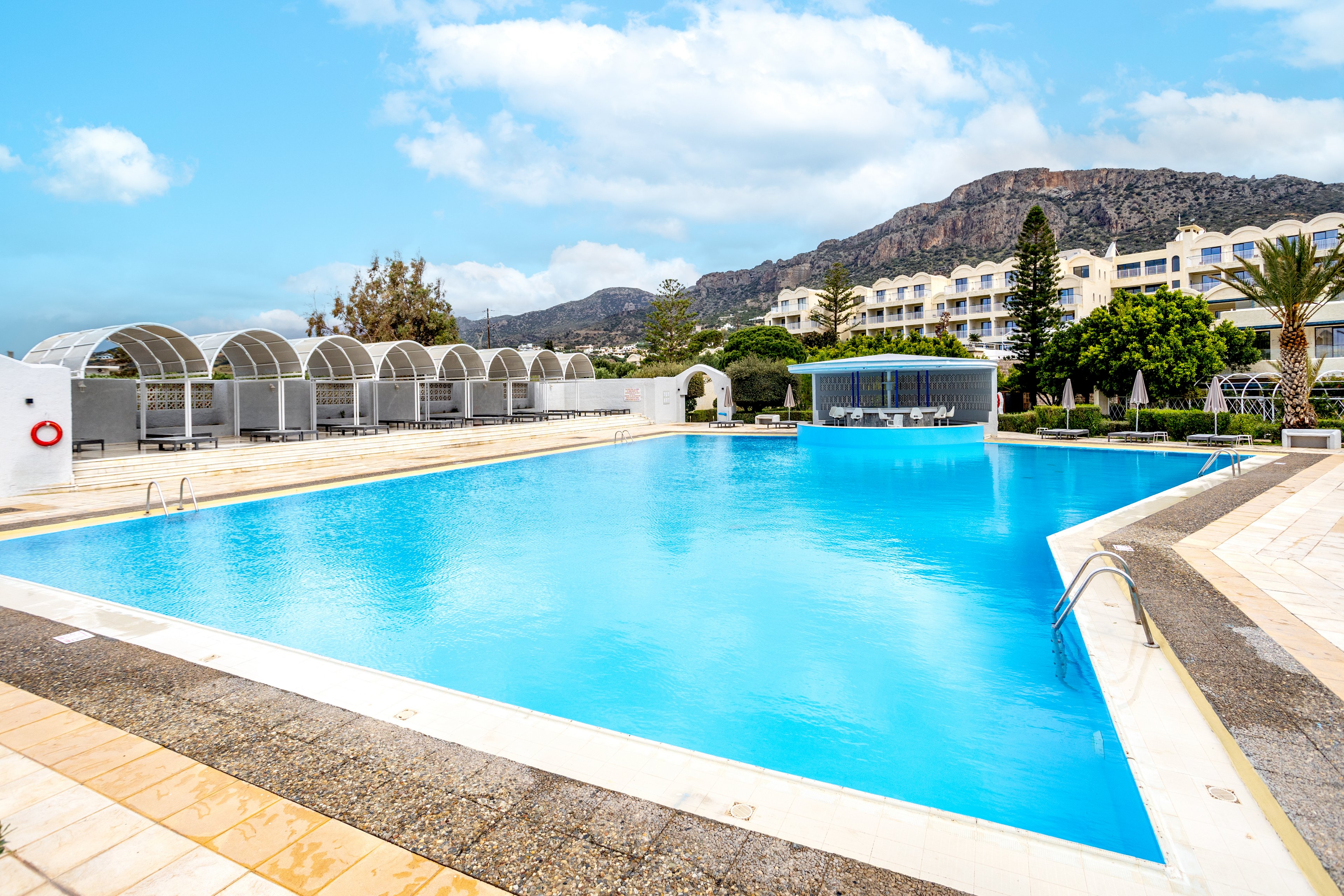 A large, clear blue swimming pool dominates the foreground of a modern resort. White poolside cabanas line one side, while a curved pool edge and metal ladders invite swimmers into the water. In the background, hotel buildings and lush greenery sit beneath a rugged mountain range. The bright sky with scattered clouds enhances the sunny, vacation atmosphere.