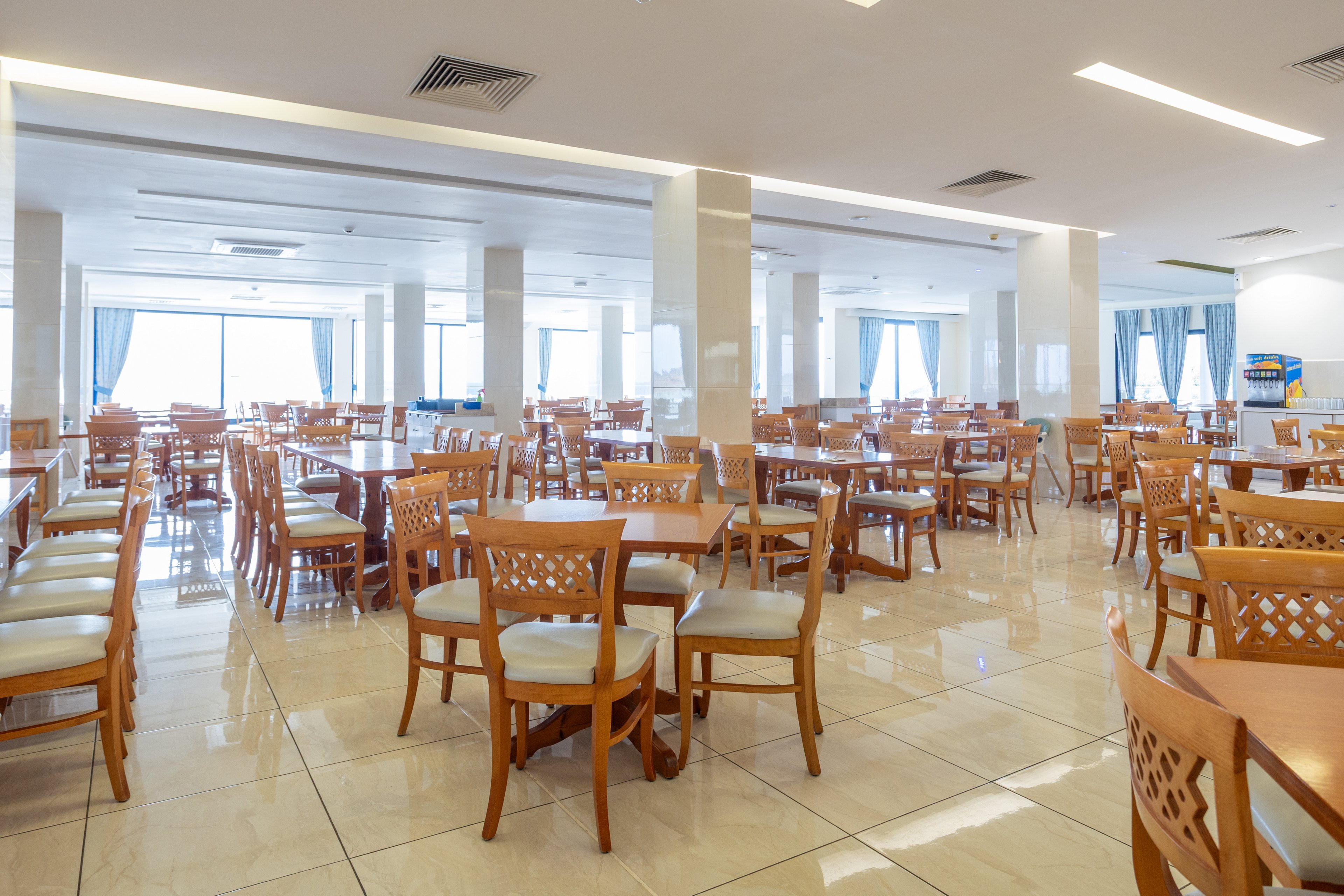 A spacious cafeteria is filled with neatly arranged wooden tables and chairs. The room is brightly lit with recessed ceiling lights and large windows at the far end. Glossy tiled floors and white columns create a clean, modern atmosphere. Blue curtains and a vending area in the background add subtle color accents to the neutral interior.