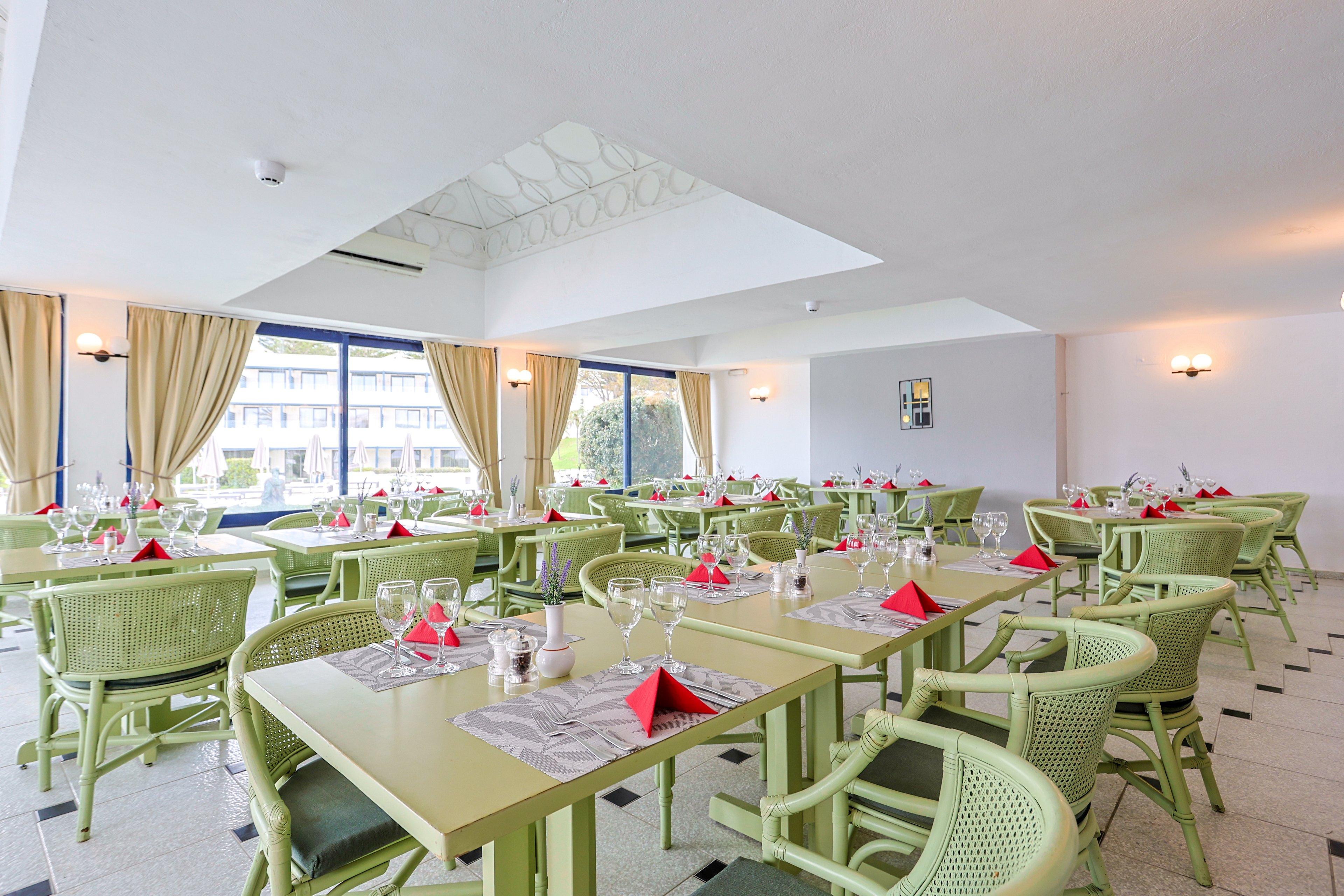 A spacious dining room is arranged with multiple pastel green tables and chairs, each neatly set with glassware and folded red napkins. Large windows along one wall let in natural light and reveal an exterior view of another building and greenery. Beige curtains, wall sconces, and a light-colored ceiling contribute to the airy, modern atmosphere. The room appears prepared for guests, suggesting a hotel restaurant or banquet space.