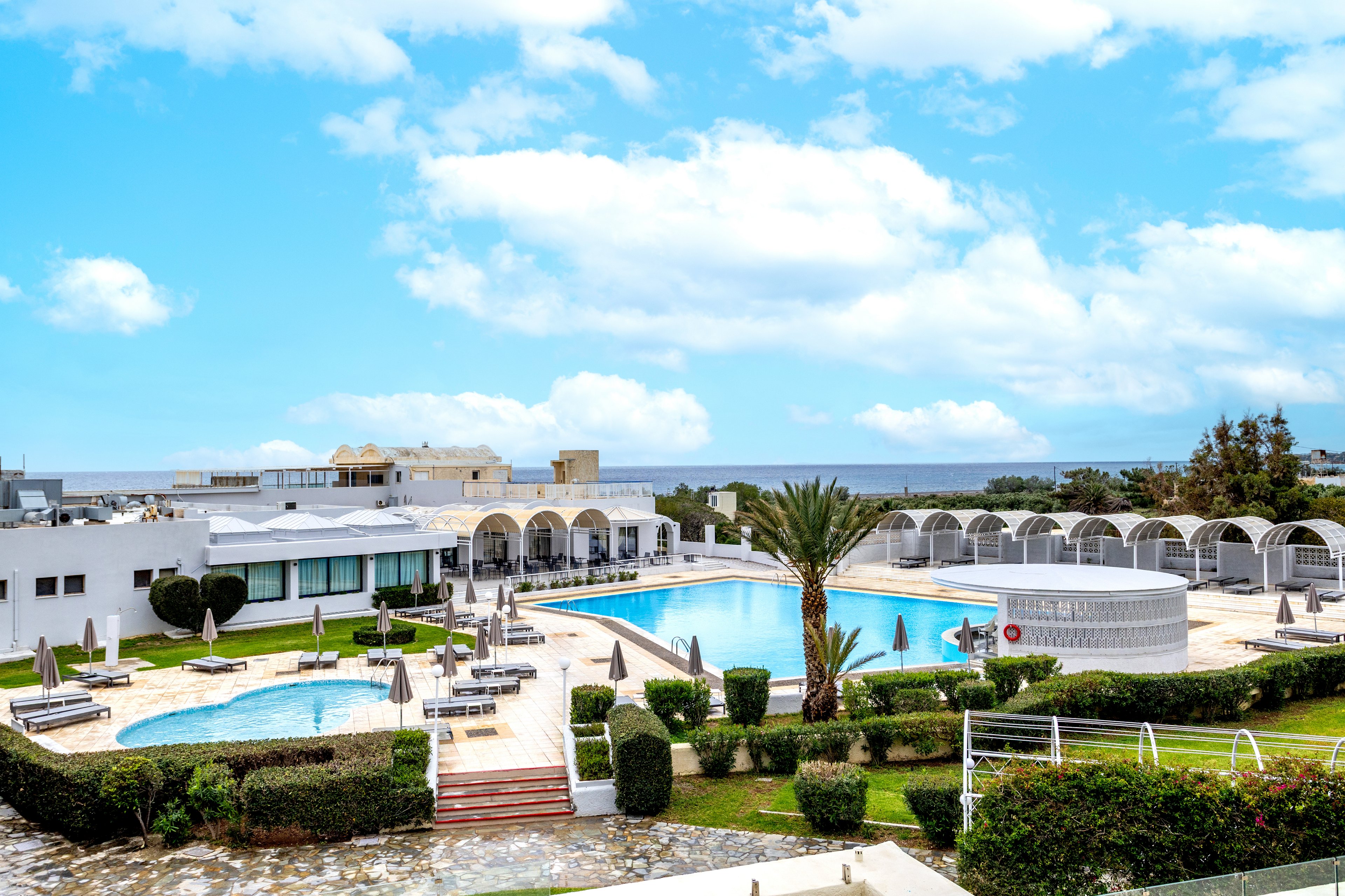 A bright seaside resort features multiple swimming pools surrounded by manicured hedges and palm trees. White low-rise buildings with arched windows and shaded lounge areas frame the pool deck. Numerous sun loungers and umbrellas are arranged neatly around the water, suggesting a relaxing vacation setting. In the distance, the ocean and a partly cloudy blue sky complete the tranquil coastal atmosphere.