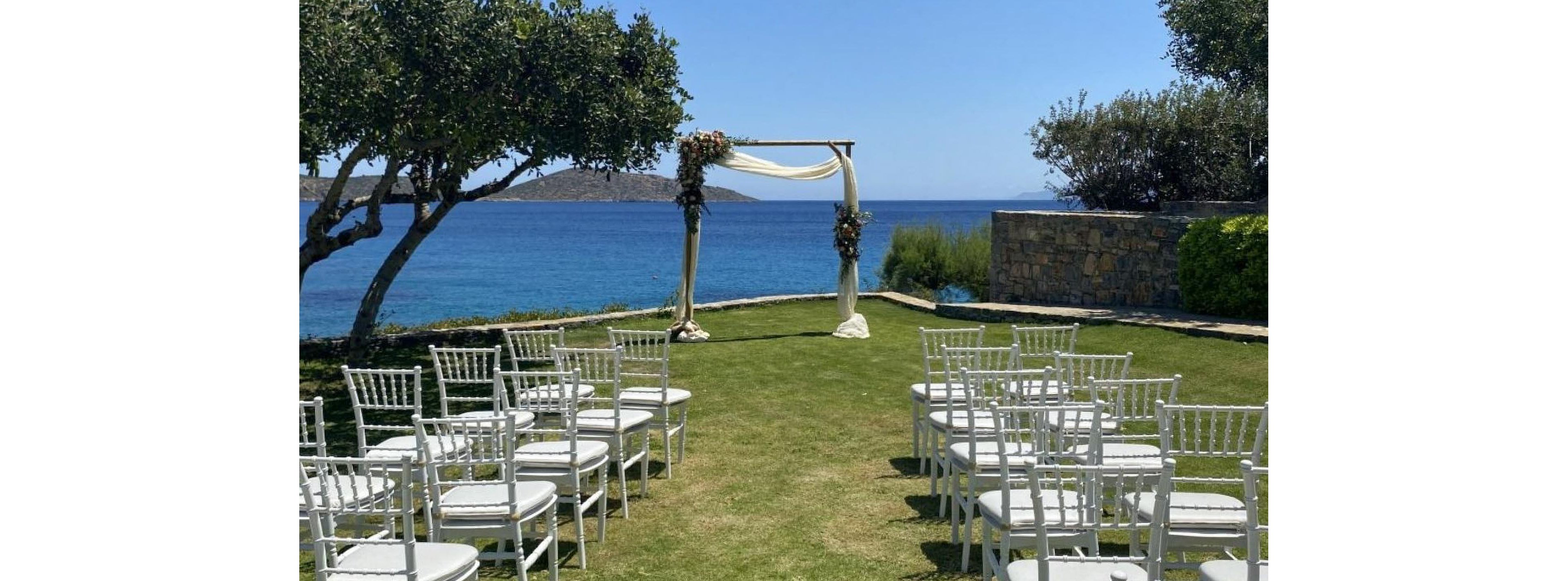 An outdoor wedding ceremony setup is arranged on a grassy lawn overlooking a calm blue sea. White chairs are lined in neat rows facing a simple wooden arch draped with light fabric and floral decorations. Olive trees frame the scene, and a stone wall borders the right side of the venue. The clear sky and bright sunlight create a serene and romantic coastal atmosphere.