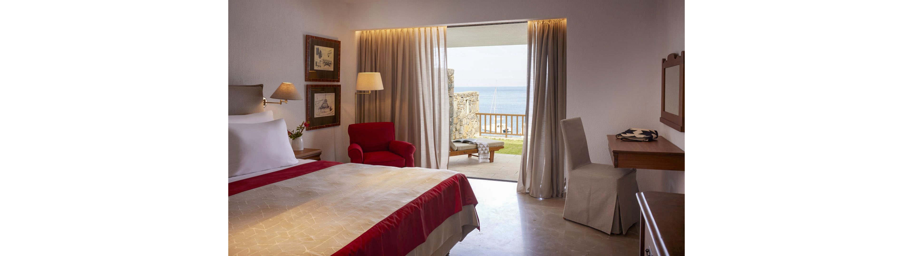 A neatly made bed with red accents dominates a cozy hotel room interior. Soft lighting, framed wall art, and a red armchair create a warm, inviting atmosphere. Floor-to-ceiling curtains open to reveal a balcony with outdoor seating and a clear view of the sea and grassy area. A simple desk and chair sit to the side, enhancing the room’s functional yet relaxing design.