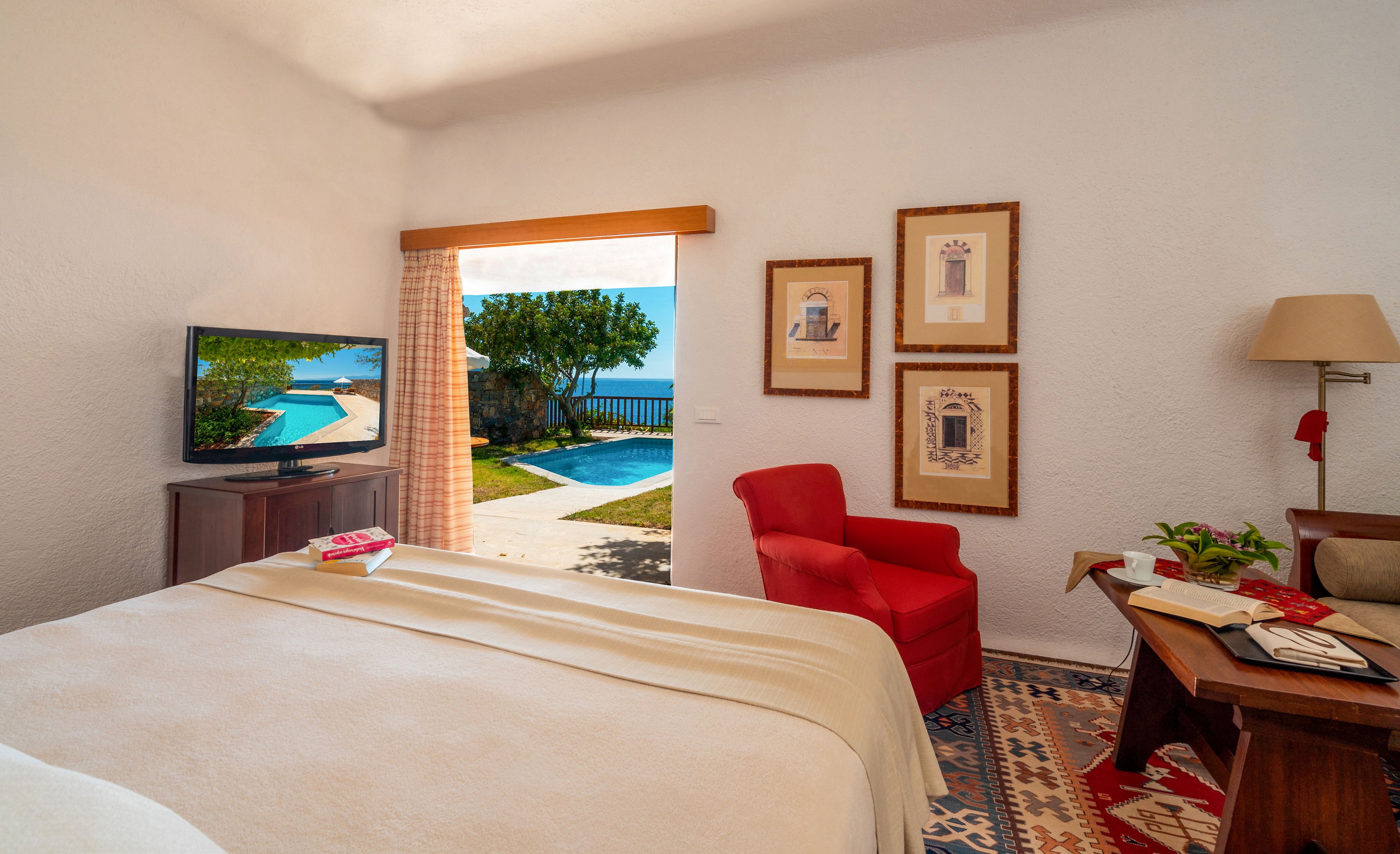 A bright, cozy hotel room features a neatly made bed facing a television and a large open doorway. Outside, a swimming pool and lush greenery are visible, suggesting a relaxing vacation setting. Inside, a red armchair, framed wall art, and a wooden desk with books and papers add warmth and character. Natural light fills the space, creating an inviting and tranquil atmosphere.