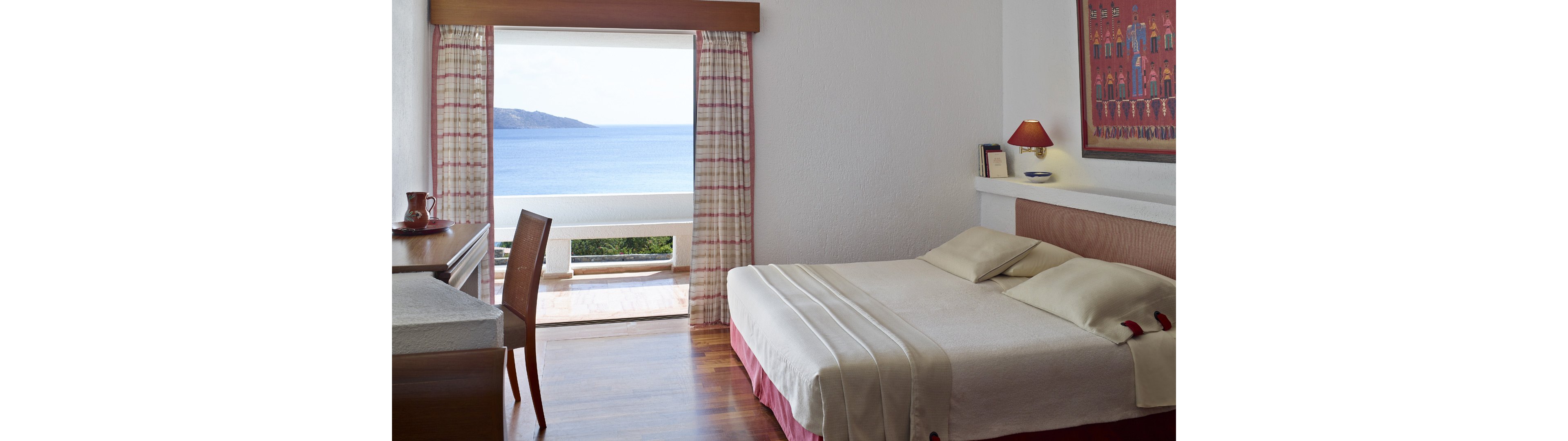 A neatly made double bed sits in a bright hotel room facing a large window with a view of the ocean. Soft neutral bedding and pillows create a calm, minimalist atmosphere. A desk and chair are positioned near the window, while a framed artwork and bedside lamp add warm accents. Sunlight streams in from the balcony area, highlighting the wooden floor and serene seaside landscape.