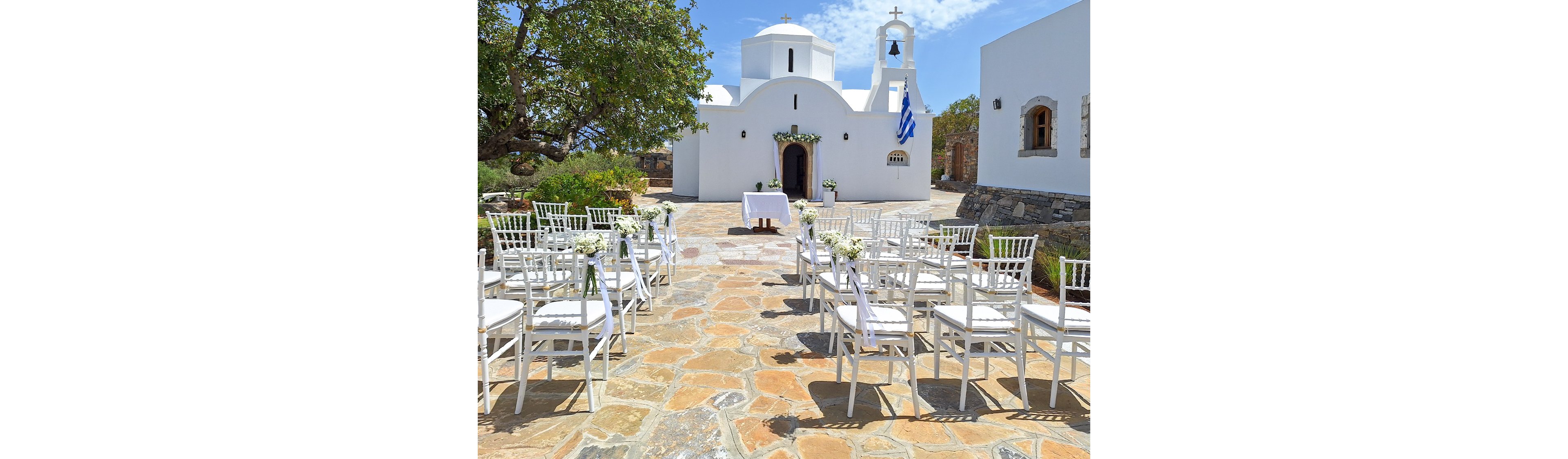 An outdoor wedding ceremony is arranged in front of a small white chapel with traditional domes. White chairs with floral decorations line a stone-paved aisle leading to a table at the entrance. The bright blue sky and sunlight emphasize the clean white architecture and warm stone tones. A flag waves beside the chapel, adding a coastal island atmosphere to the serene scene.