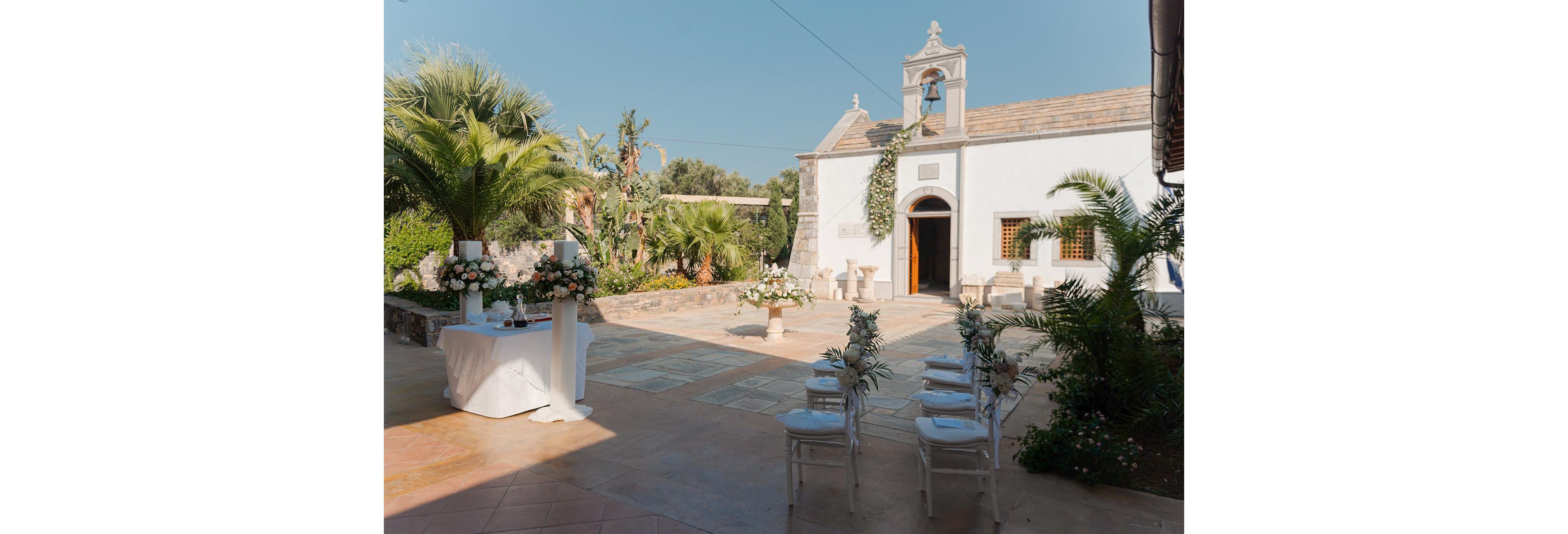 An intimate outdoor wedding ceremony is set up in front of a small white chapel with an open wooden door. White chairs decorated with greenery and flowers face a central floral arrangement in the courtyard. A table with elegant white linens and flower centerpieces stands to the side, surrounded by palm trees and lush garden plants. The scene is bathed in warm daylight, creating a serene and romantic atmosphere.