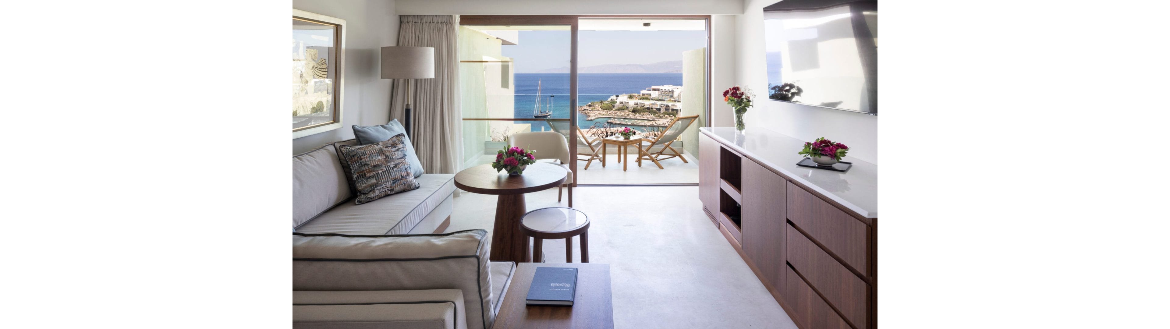 A contemporary hotel suite features a light-colored sofa, wooden furniture, and a sleek built-in cabinet. Large sliding glass doors open onto a balcony with chairs and a small table overlooking the sea. The view includes a marina with boats and white buildings along the coastline. Bright natural light and neutral tones create an airy, relaxing atmosphere ideal for a beach vacation.