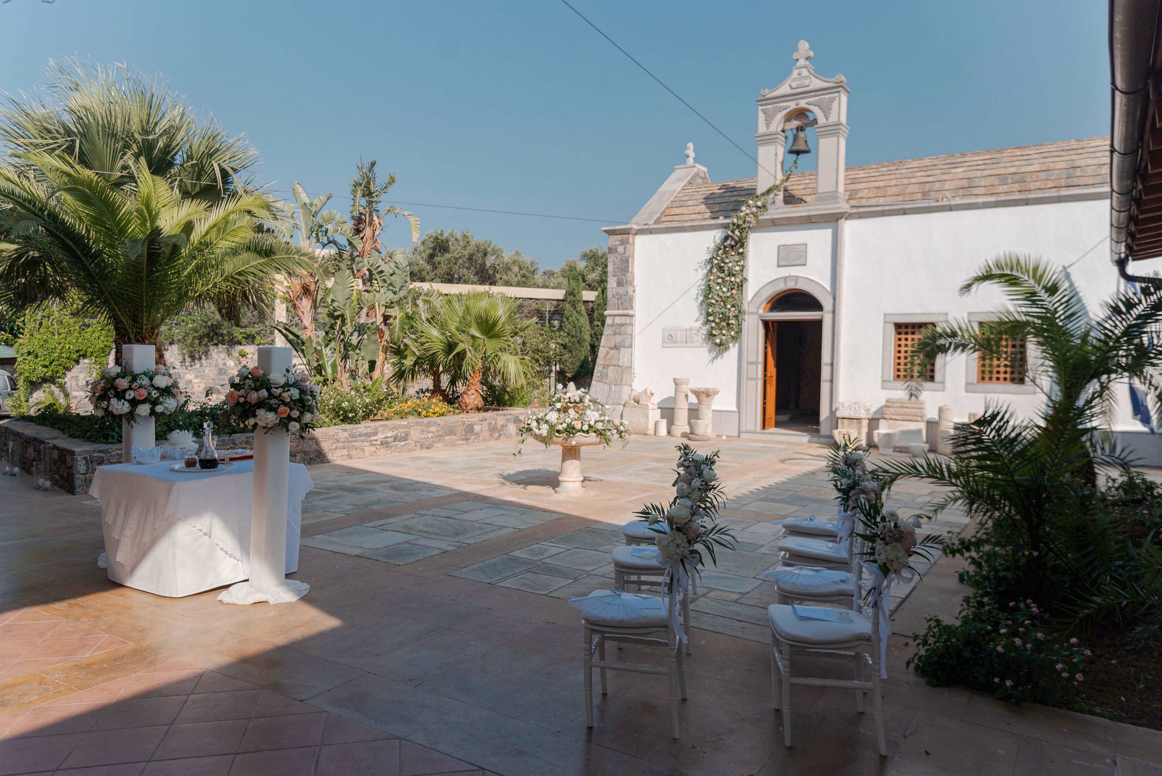 An intimate outdoor wedding ceremony is arranged in front of a small white chapel with an open wooden door. White chairs decorated with floral arrangements are lined up facing a central aisle and altar area. Lush green palm trees and garden plants surround the stone courtyard, creating a serene Mediterranean atmosphere. Soft daylight and pastel floral decorations contribute to a romantic and elegant setting.